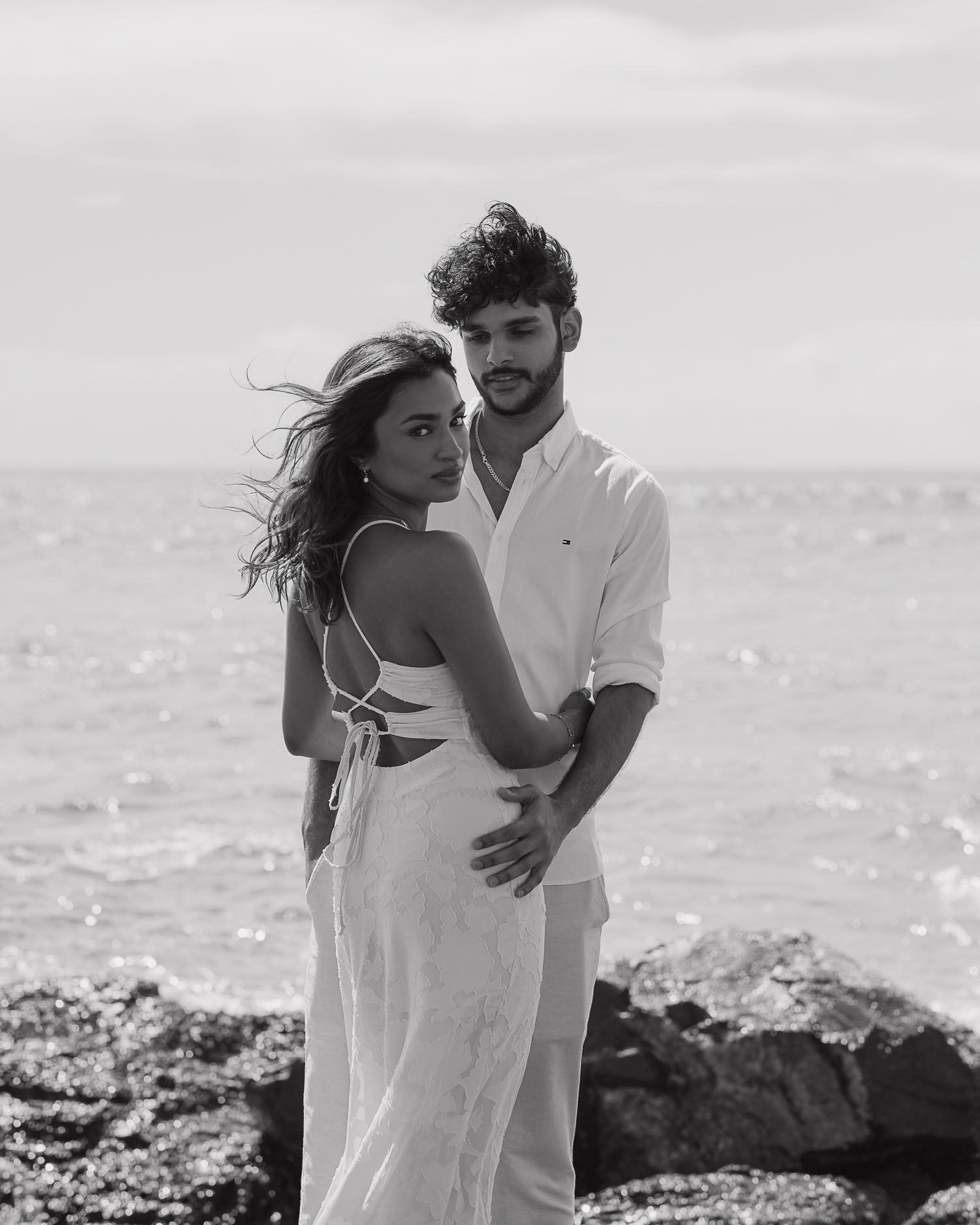 A romantic couple in white clothing shares an intimate moment by the ocean.