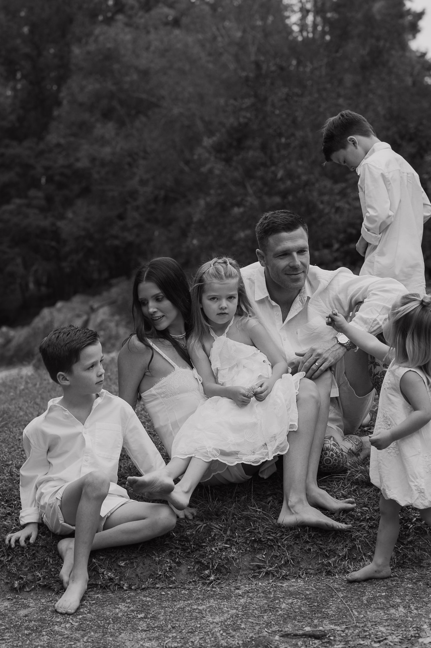 Black and white family portrait of parents with four children sitting outdoors near trees in matching white outfits.