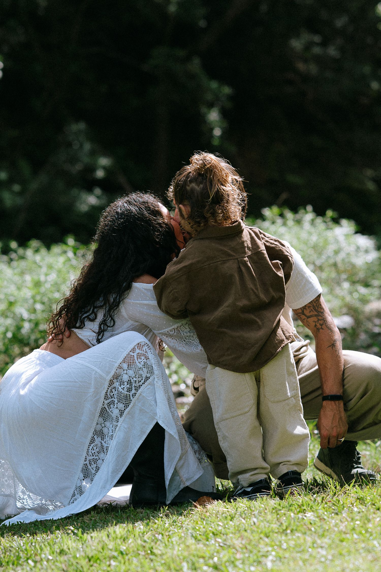 A loving family moment as a mother in a white lace dress and tattooed father embrace their young child on grass.