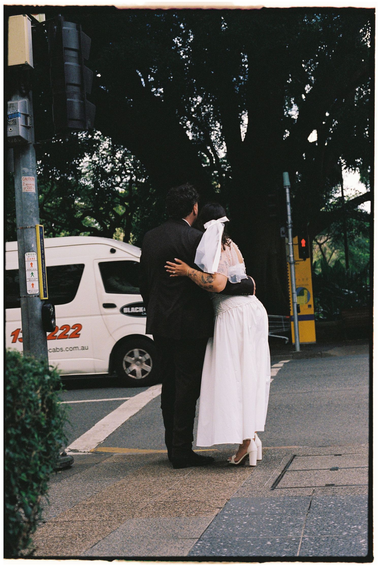 Bride and groom embracing on a city sidewalk, sharing a romantic moment in wedding attire near lush trees.