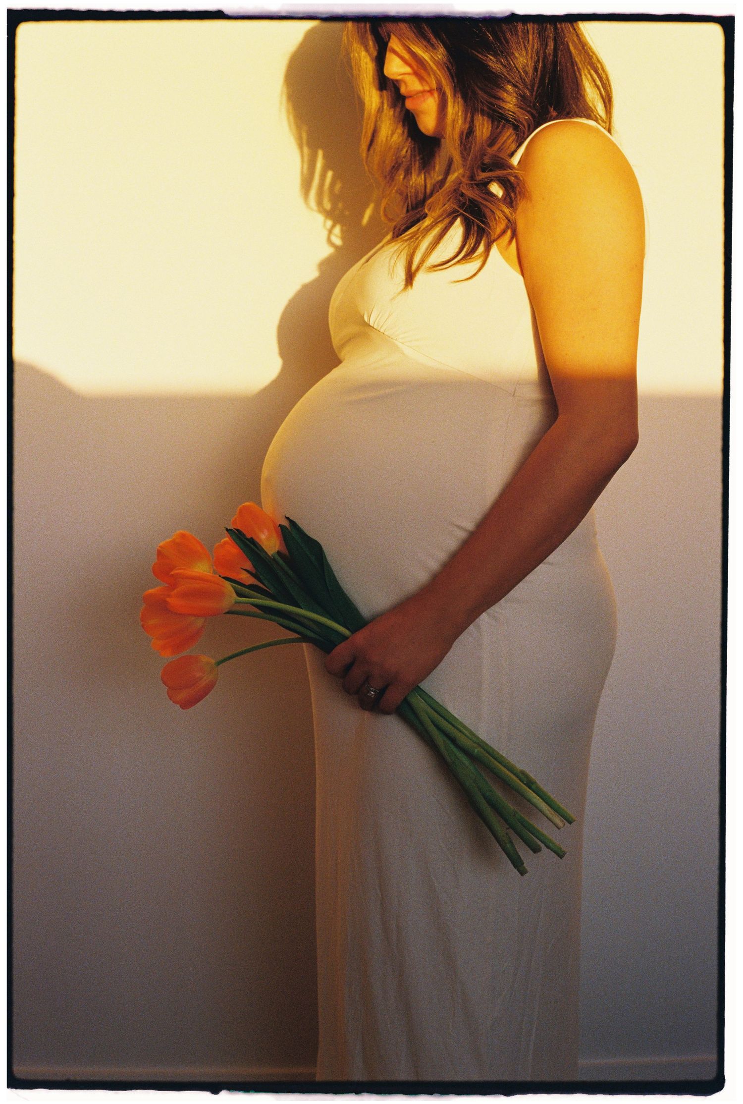 Silhouette holding orange tulips against a sunlit wall in a flowing white dress.