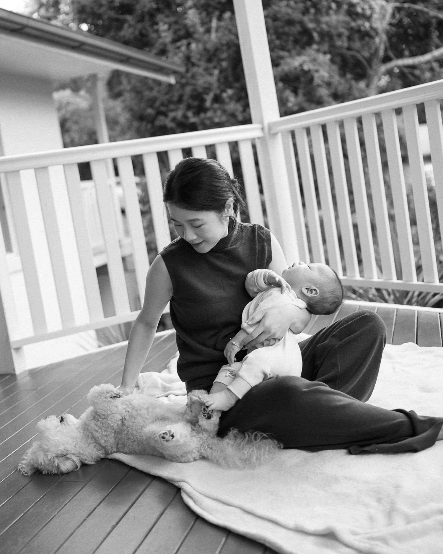 Black and white photo of a woman sitting on a deck, cradling a newborn baby with a fluffy dog lying nearby.