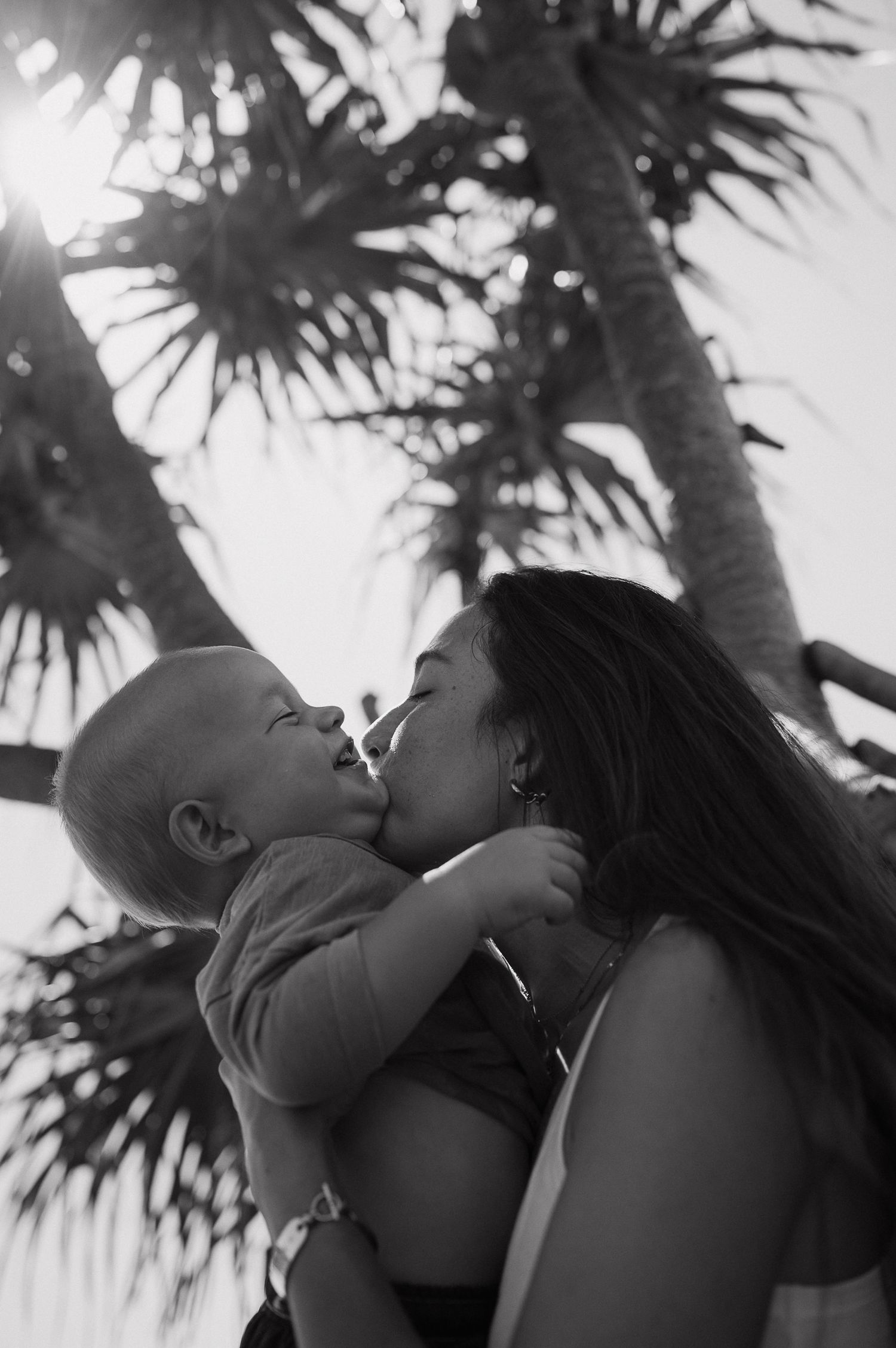 Black and white photo of a mother kissing her baby outdoors with palm trees in the background.