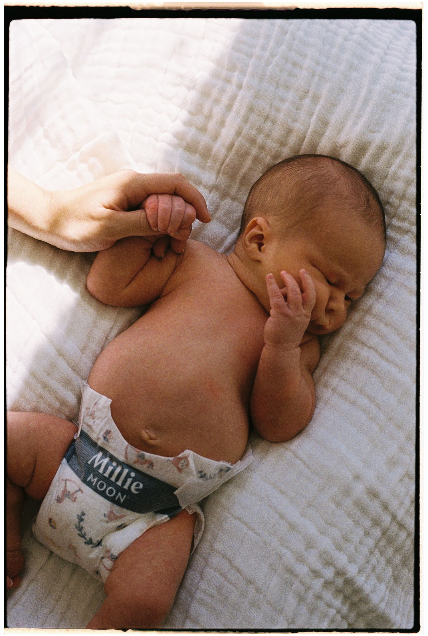 Newborn baby wearing a Millie Moon diaper lying on a white blanket while an adult gently holds their hand.