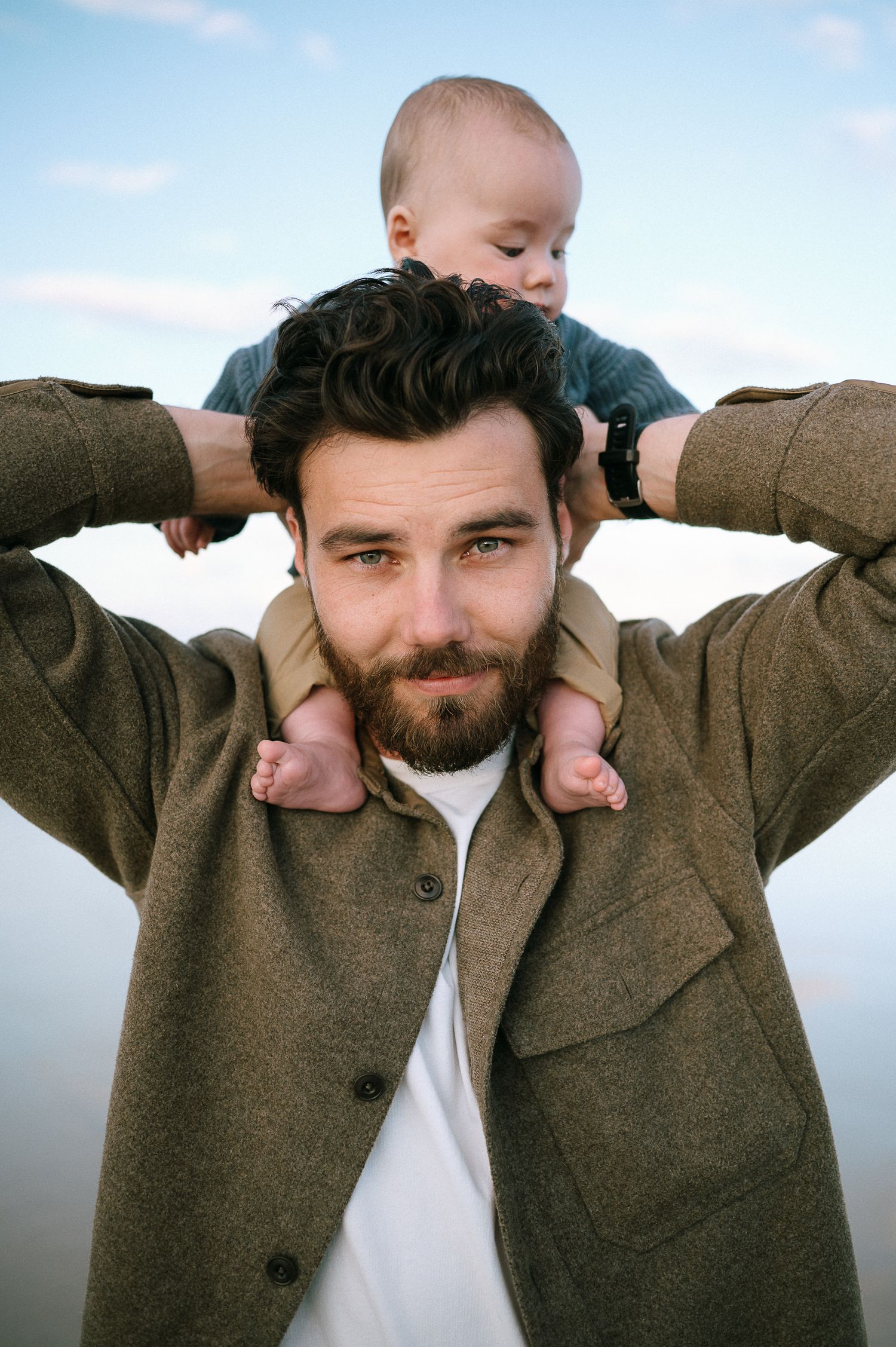 Baby sits on shoulders wearing blue sweater against cloudy sky.
