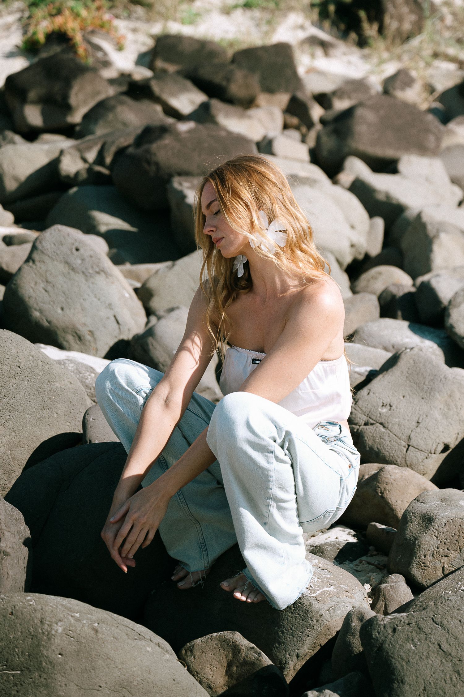Blonde woman in white top and light jeans sitting on dark rocks outdoors in sunlight.