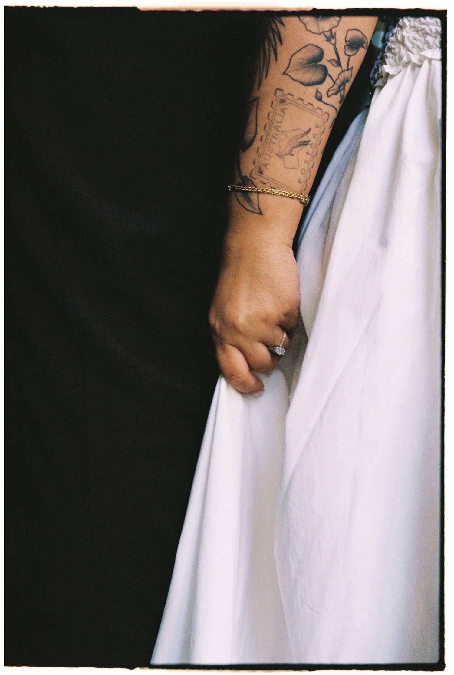 Close-up of tattooed arm with gold bracelet and diamond ring holding white wedding dress.