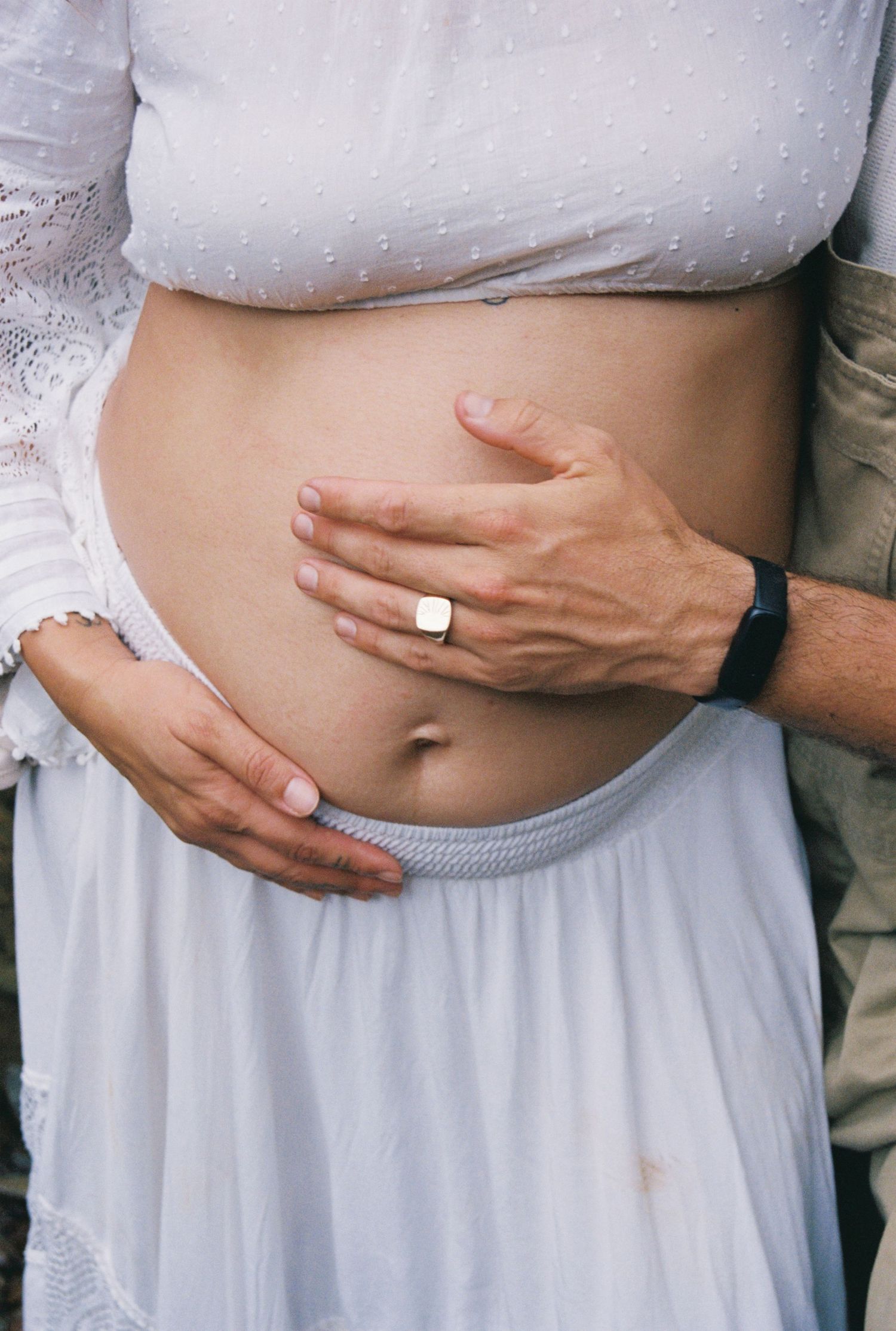 Close-up of a pregnant woman's bare baby bump being gently held by two hands, wearing a white skirt and top.
