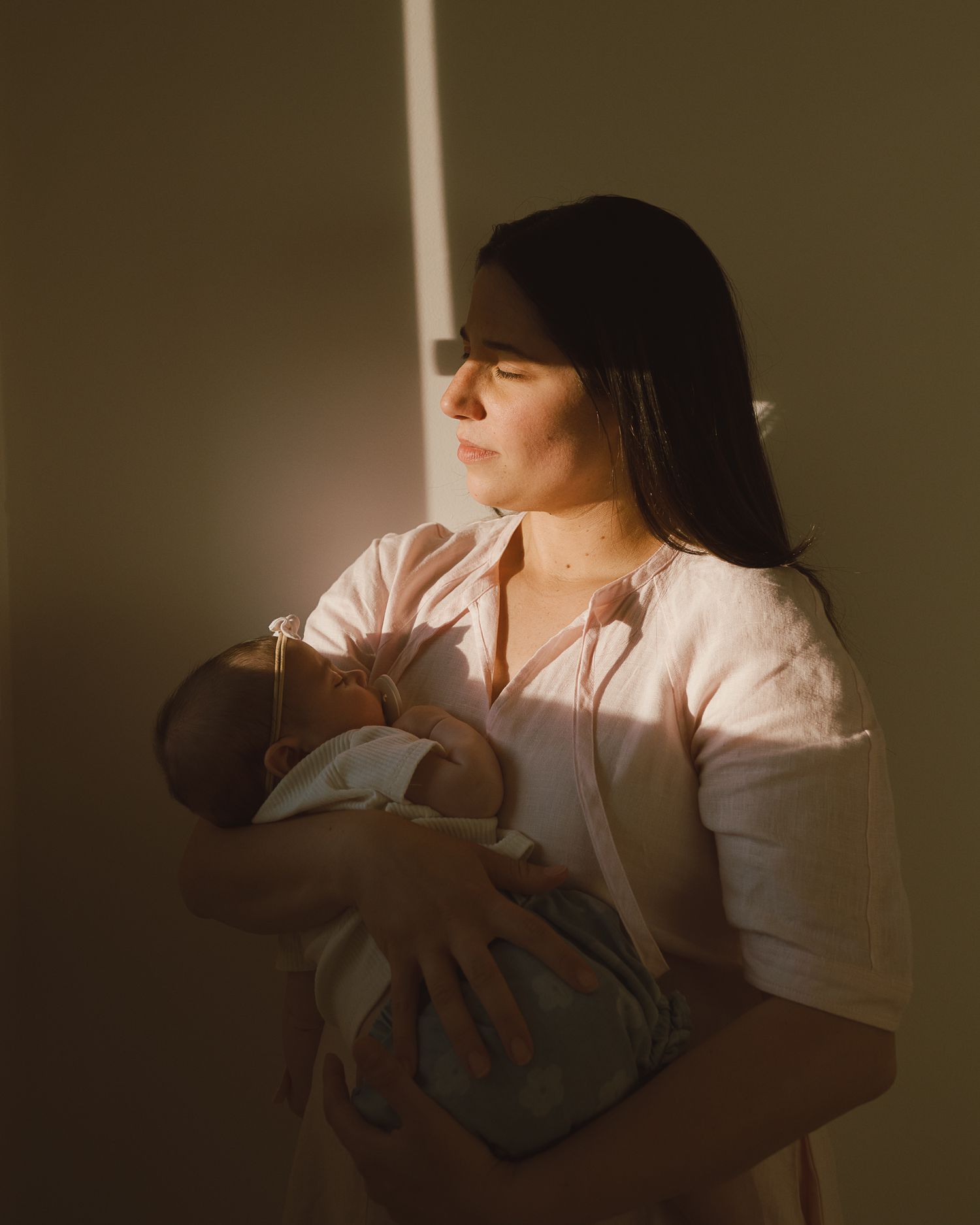 A mother tenderly holds her newborn baby in warm golden light streaming through a window.