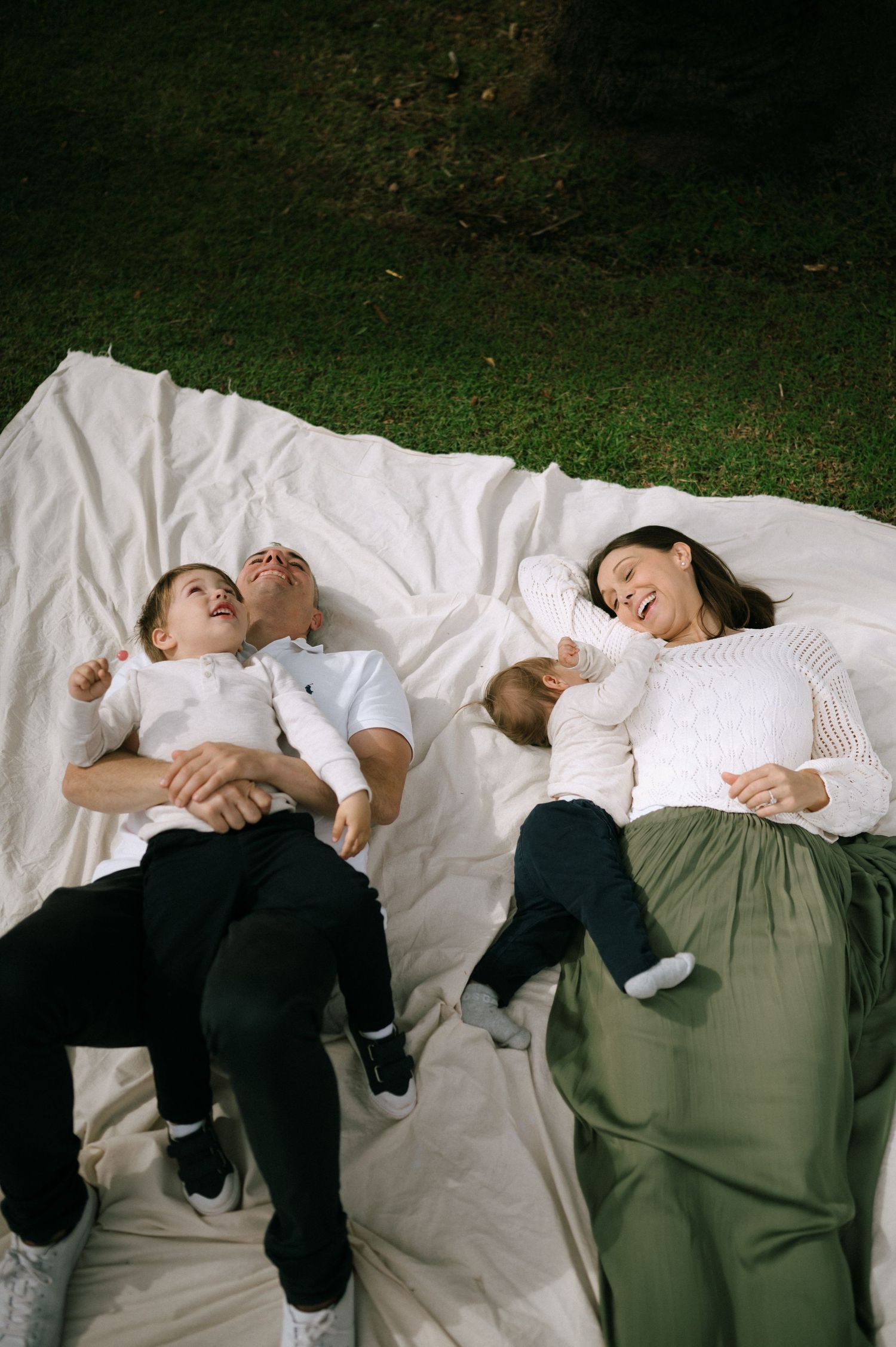 Family lying together on a white blanket in the grass.