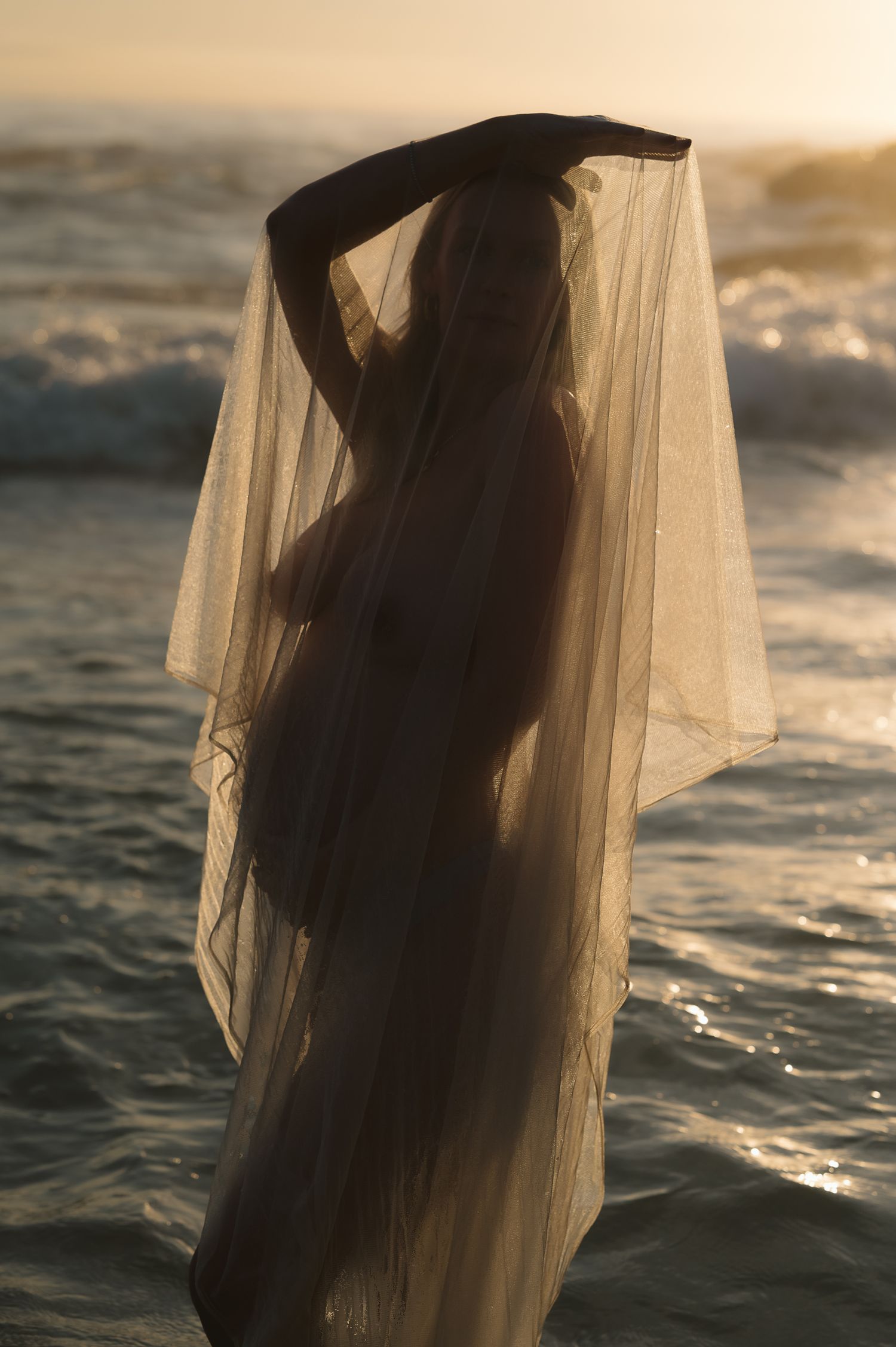 Ethereal silhouette draped in sheer fabric against sunset ocean waves.