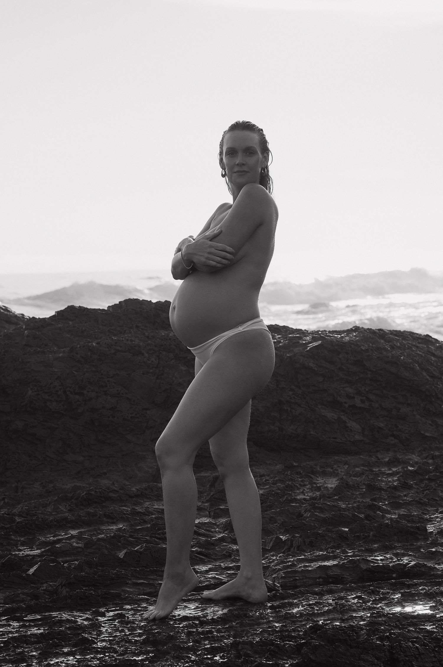 Silhouette of pregnant figure standing on rocky coastline at sunset.
