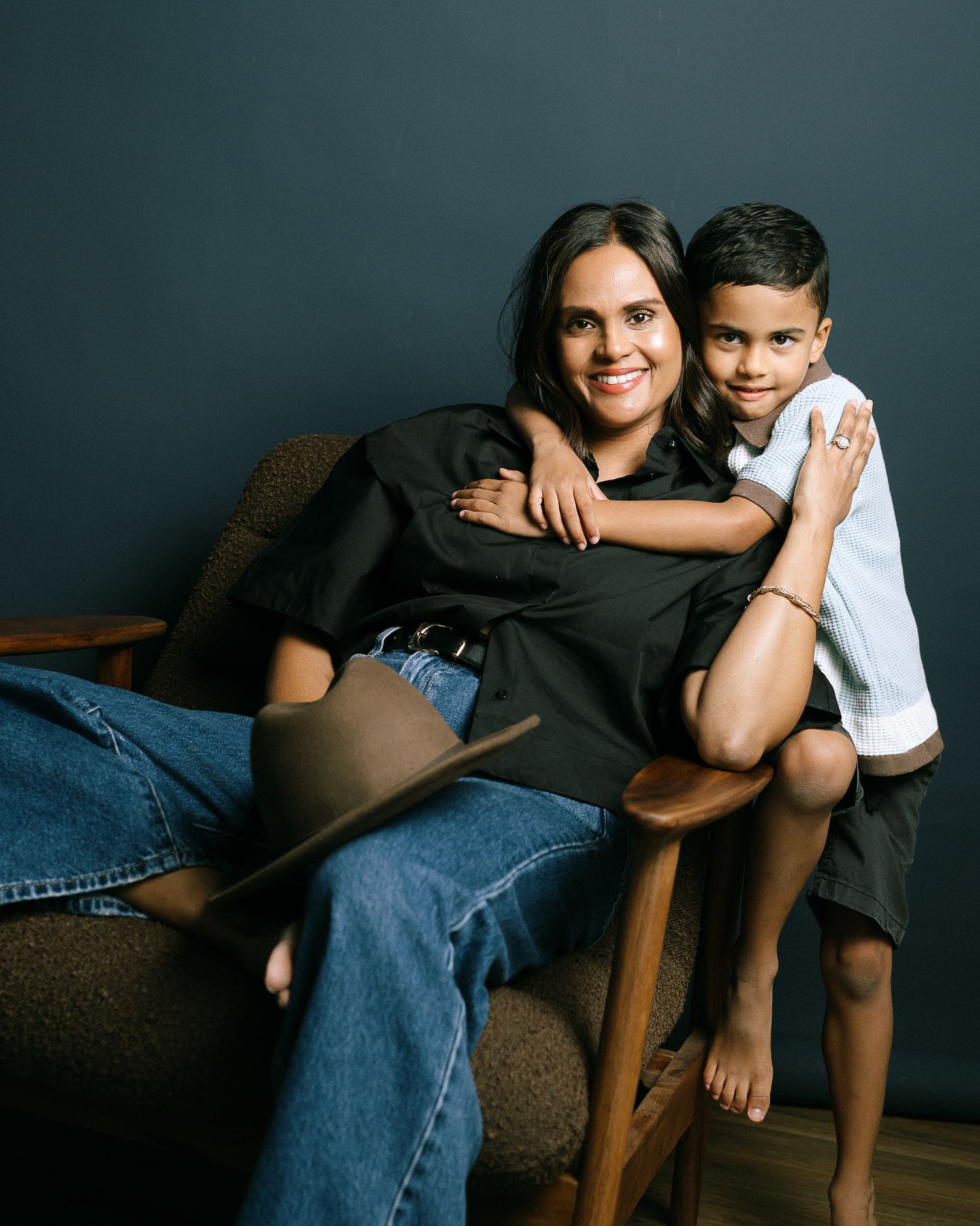 A woman in a black top and jeans sits in a chair smiling, with a young boy in a white shirt perched beside her.