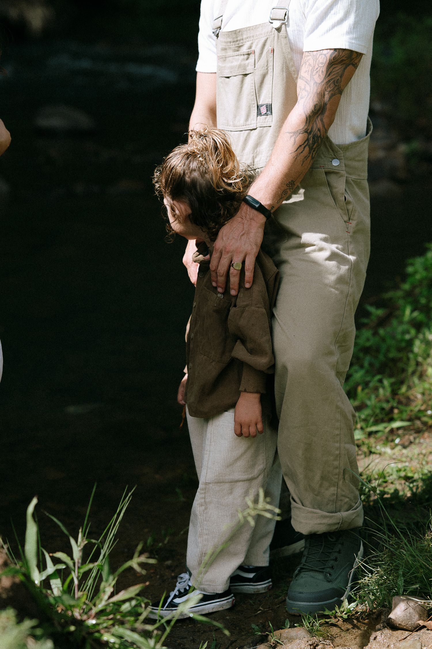 A tattooed father tenderly holds his young child close near a sunlit creek surrounded by lush greenery.
