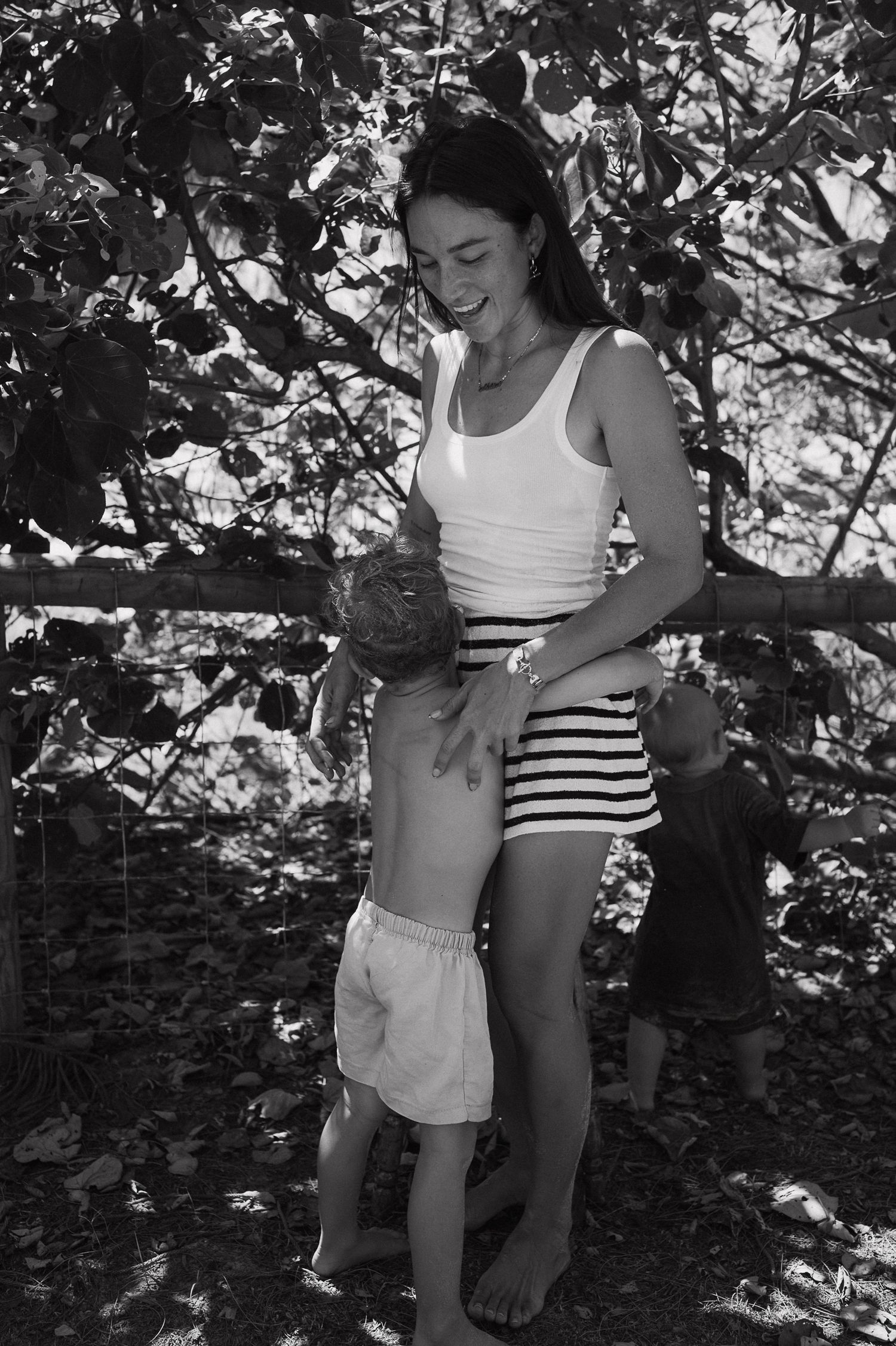 Black and white photo of a smiling woman in striped shorts holding a toddler under shady trees.