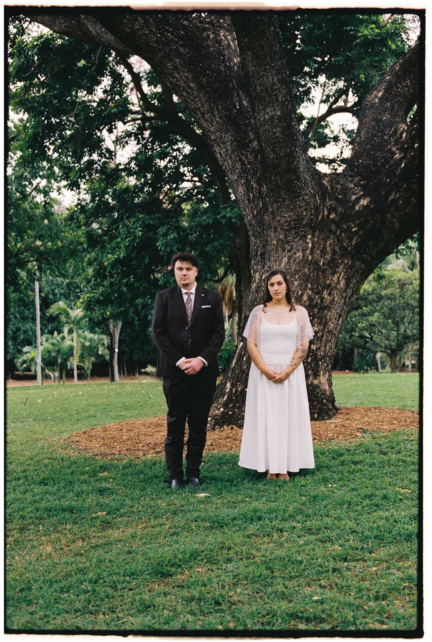 A couple in formal attire stands before a large tree on green grass, likely at an outdoor wedding ceremony.