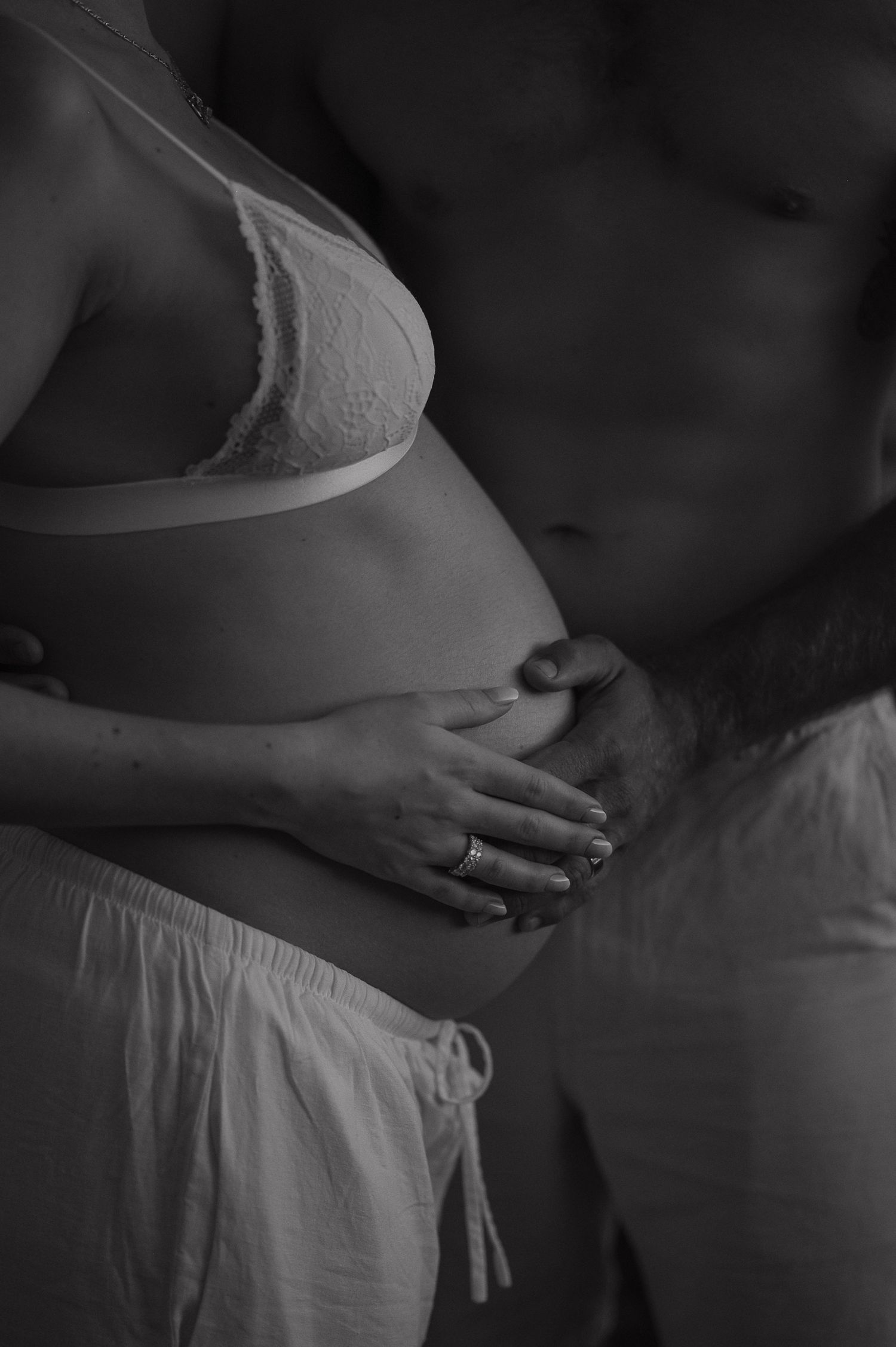 Black and white maternity photo of pregnant woman in white bra cradling baby bump with partner standing behind her.