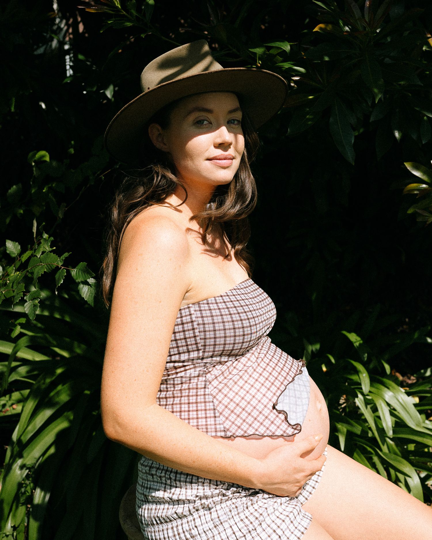 A sun-drenched maternity portrait featuring a woman in a checkered dress and wide-brimmed hat among foliage.