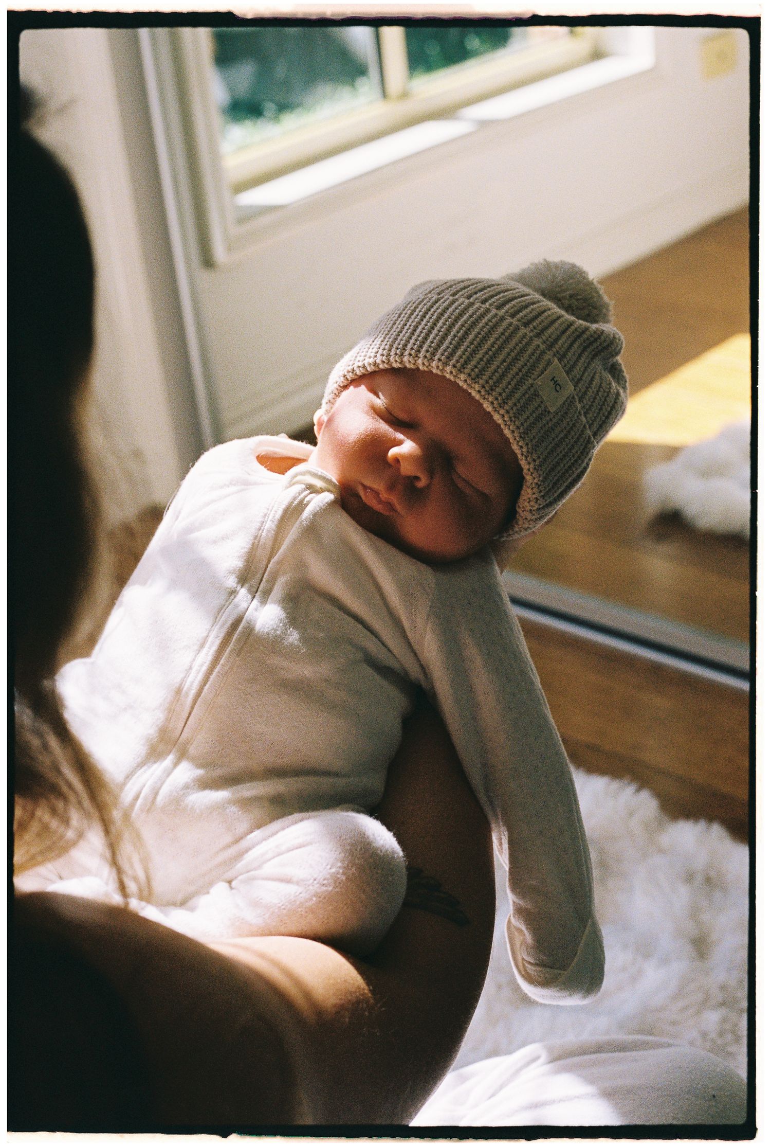 Newborn baby wearing a gray knit beanie hat, swaddled in white, sleeping peacefully in sunlit arms.