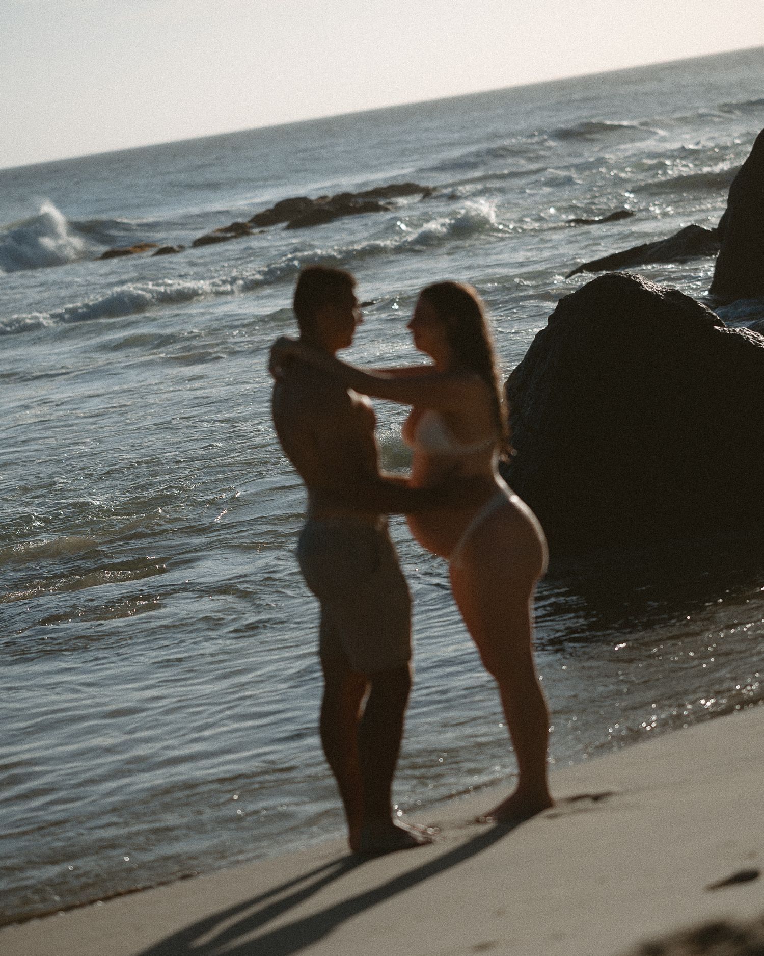 Couple embracing on a sandy beach at sunset with ocean waves and rocky shoreline in the background.