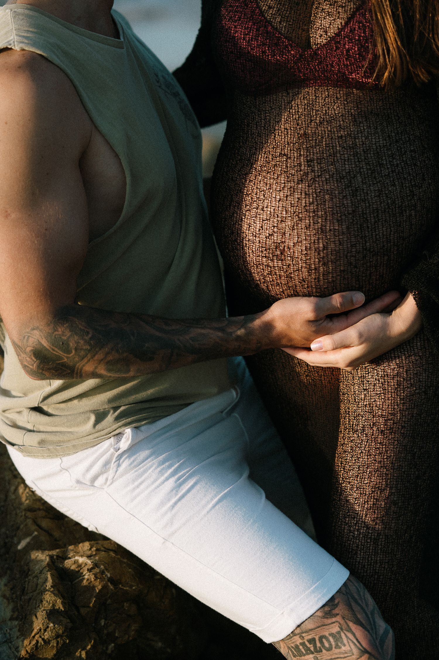 Tattooed partner tenderly holds pregnant woman's baby bump during an outdoor maternity photo session.