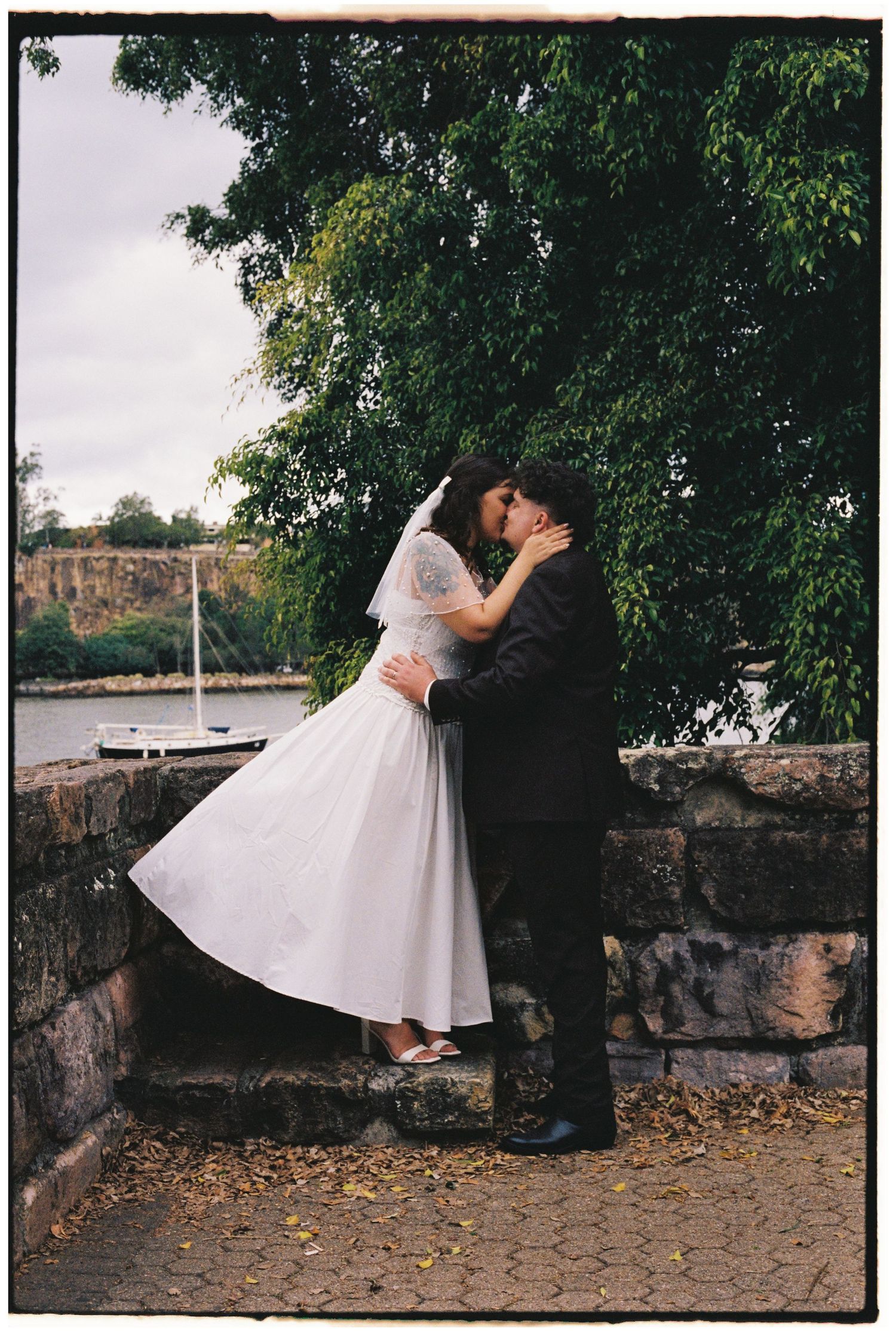 Bride and groom share romantic kiss near stone wall by waterfront, bride in tea-length white dress with veil.
