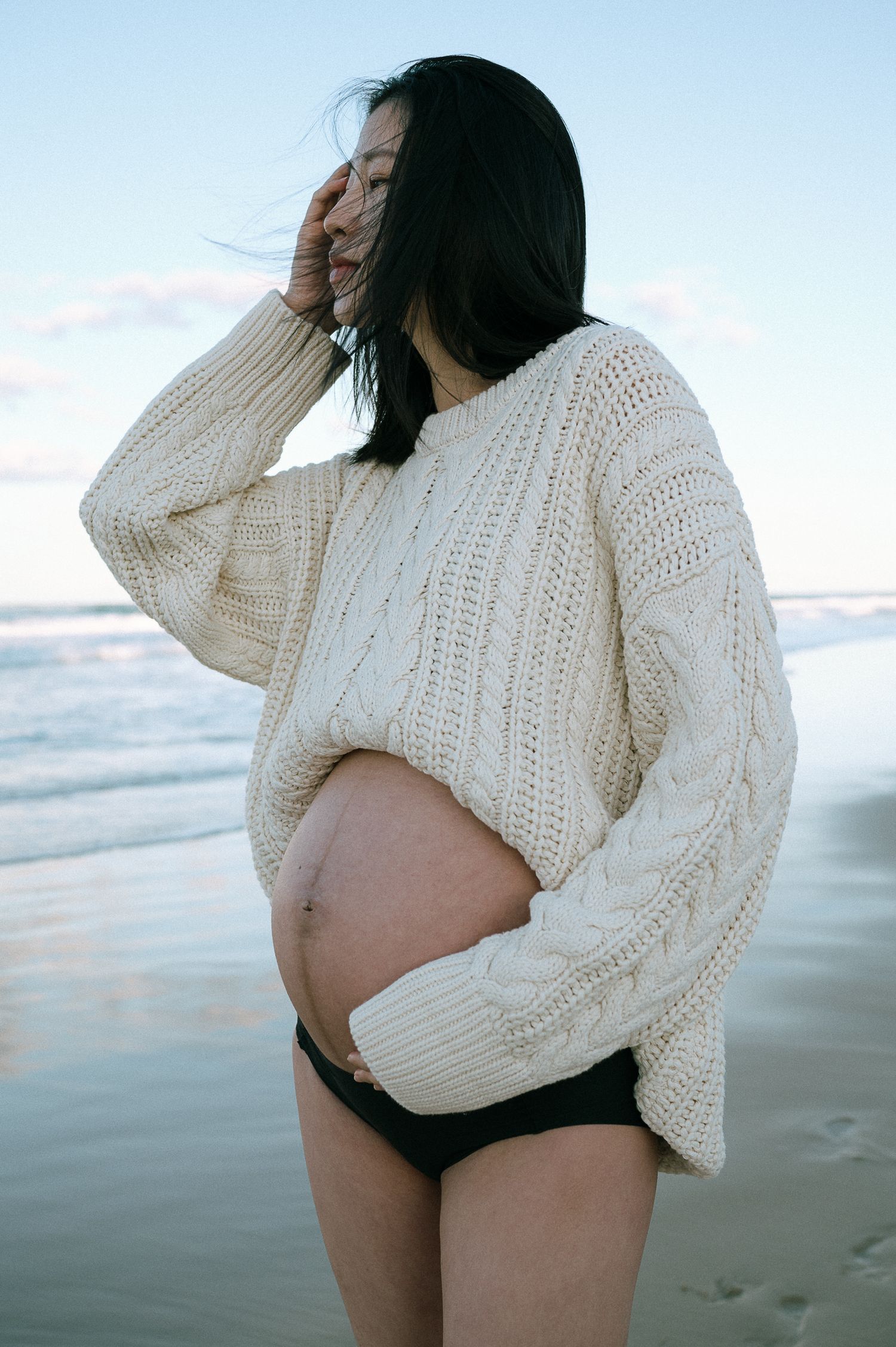 Pregnant woman in cream sweater poses by ocean during sunset.