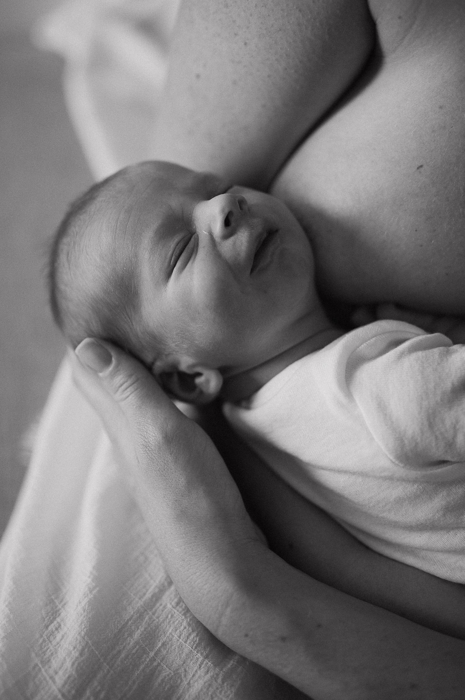 Black and white photo of a peaceful newborn baby swaddled in white, cradled gently in a parent's arms.