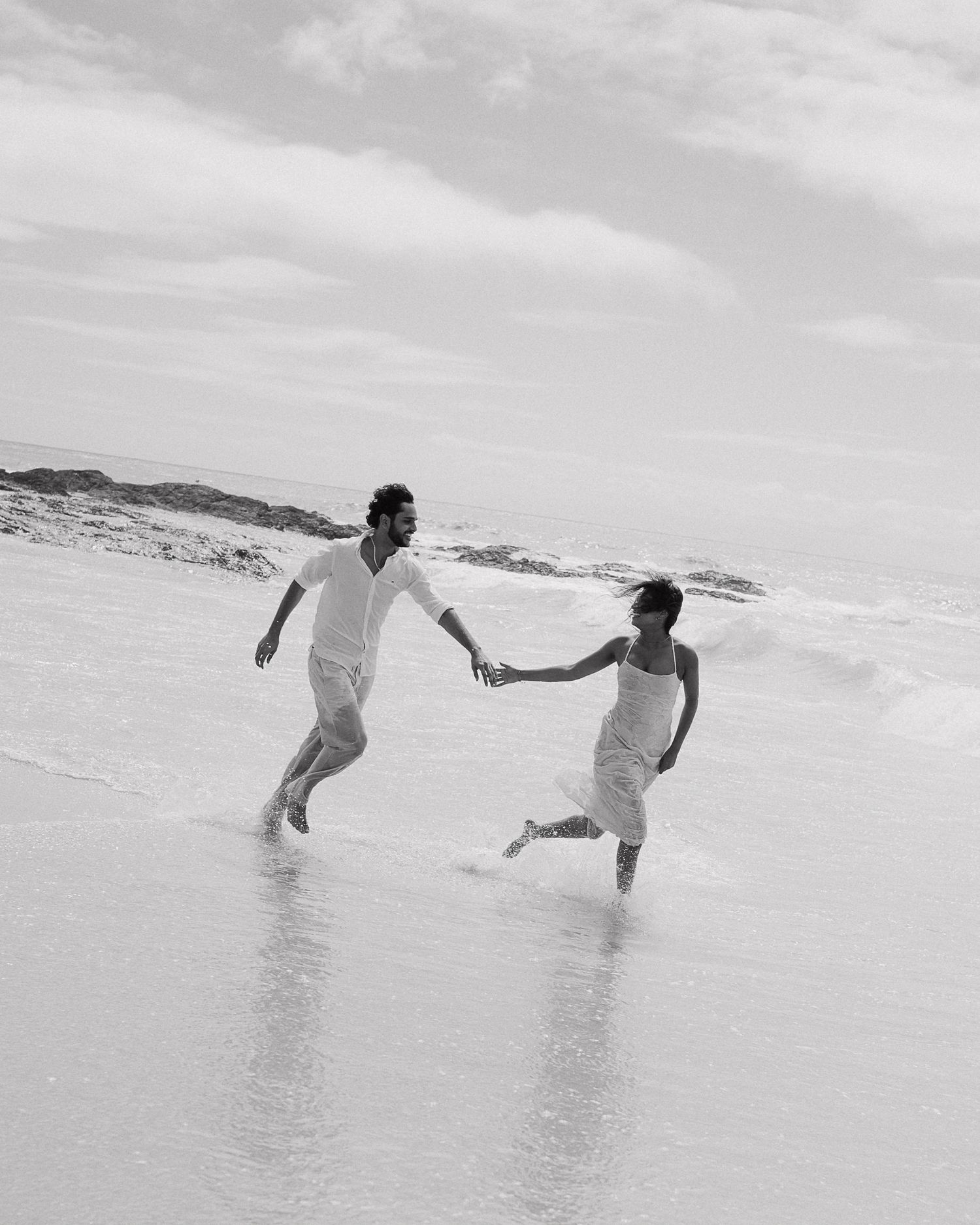 A joyful couple runs hand-in-hand through ocean waves on a beach in a black and white romantic photo.