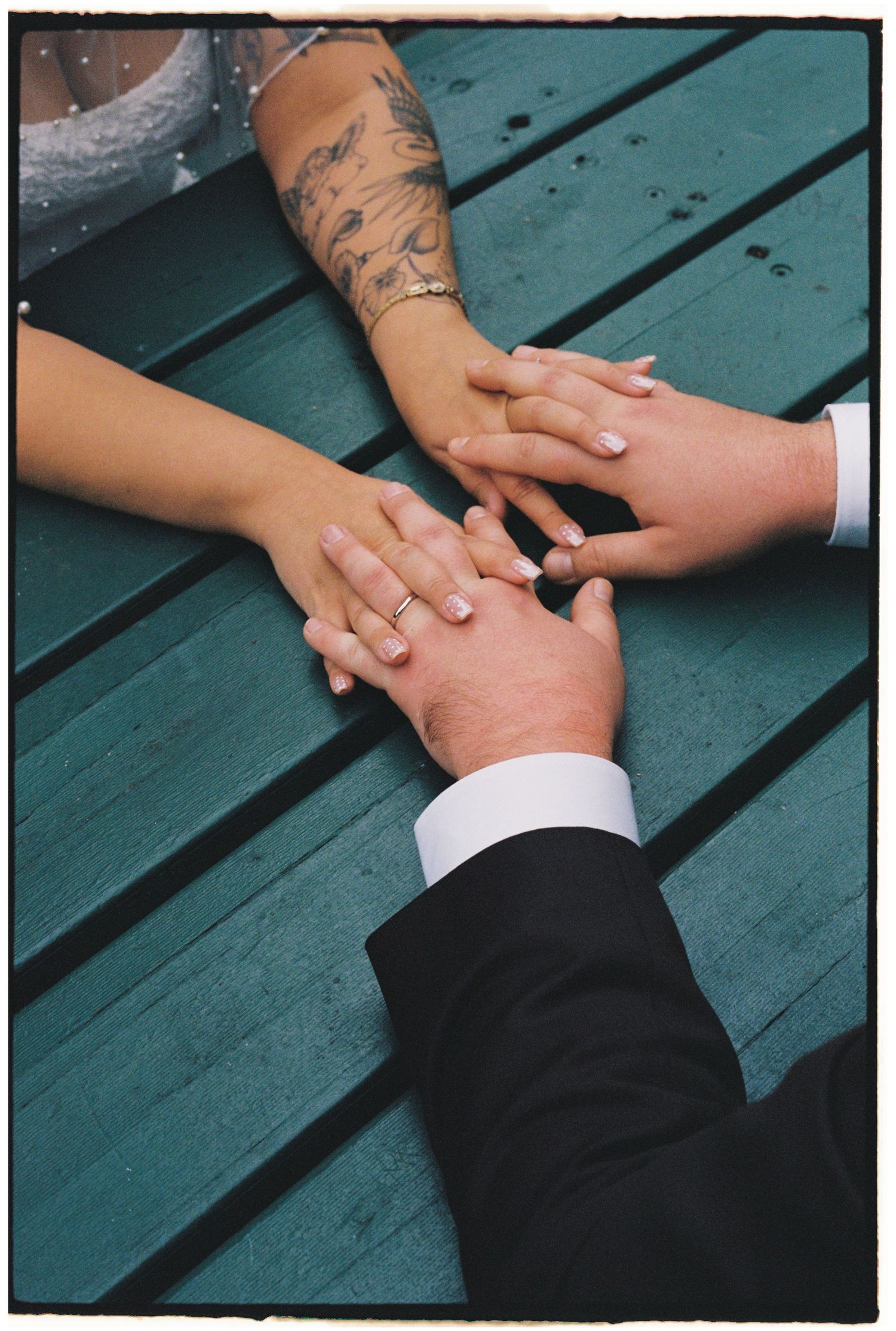 Three hands stacked together on a teal bench, featuring a wedding ring, tattoo, and formal suit sleeve.