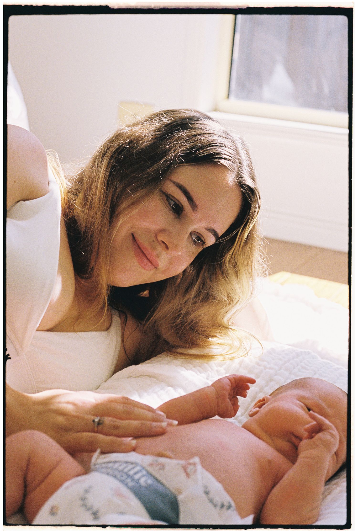 A young mother smiles tenderly at her newborn baby lying on a white blanket in soft natural light.