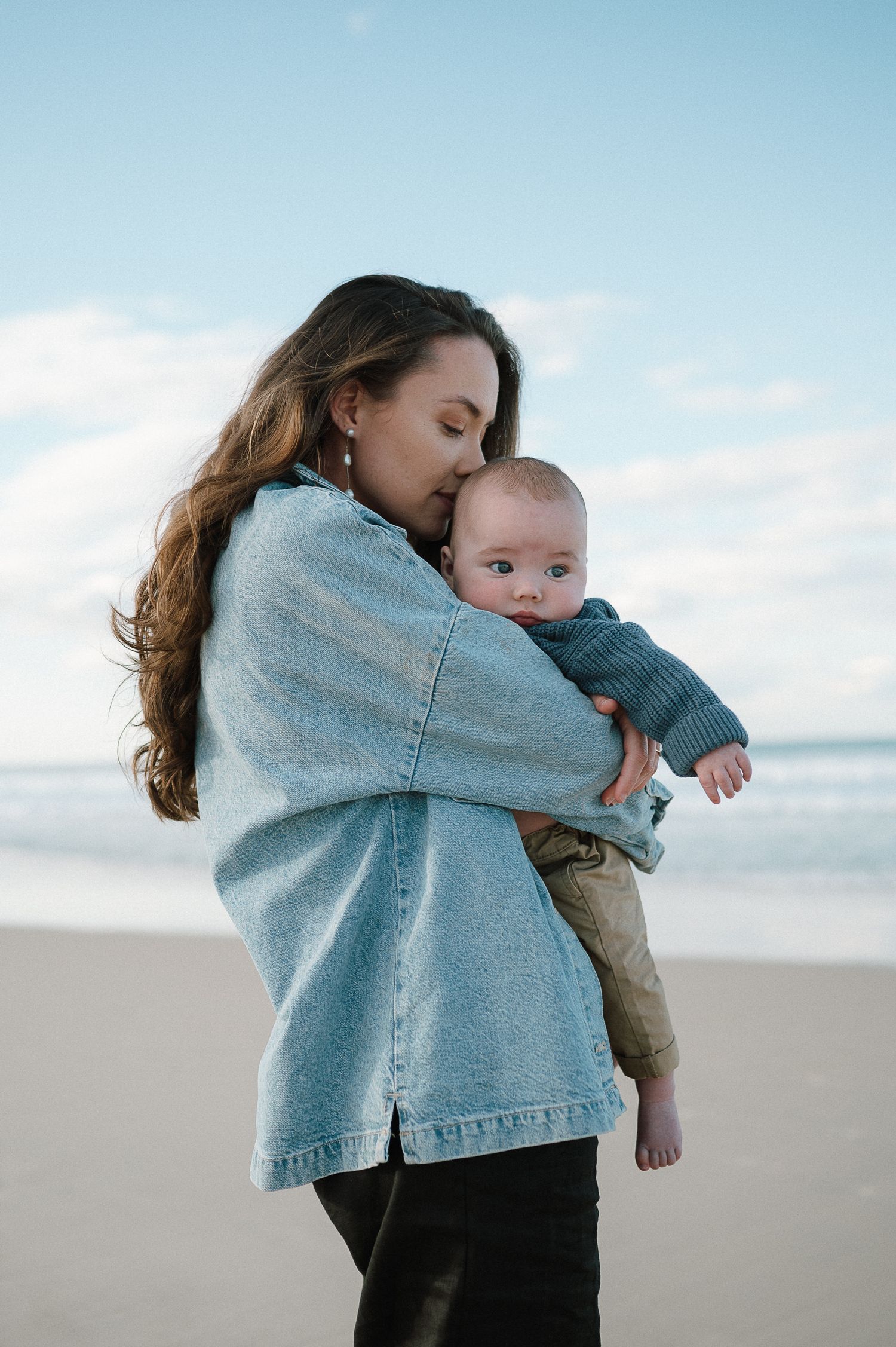 Parent embracing baby against bright beach landscape in denim jacket.