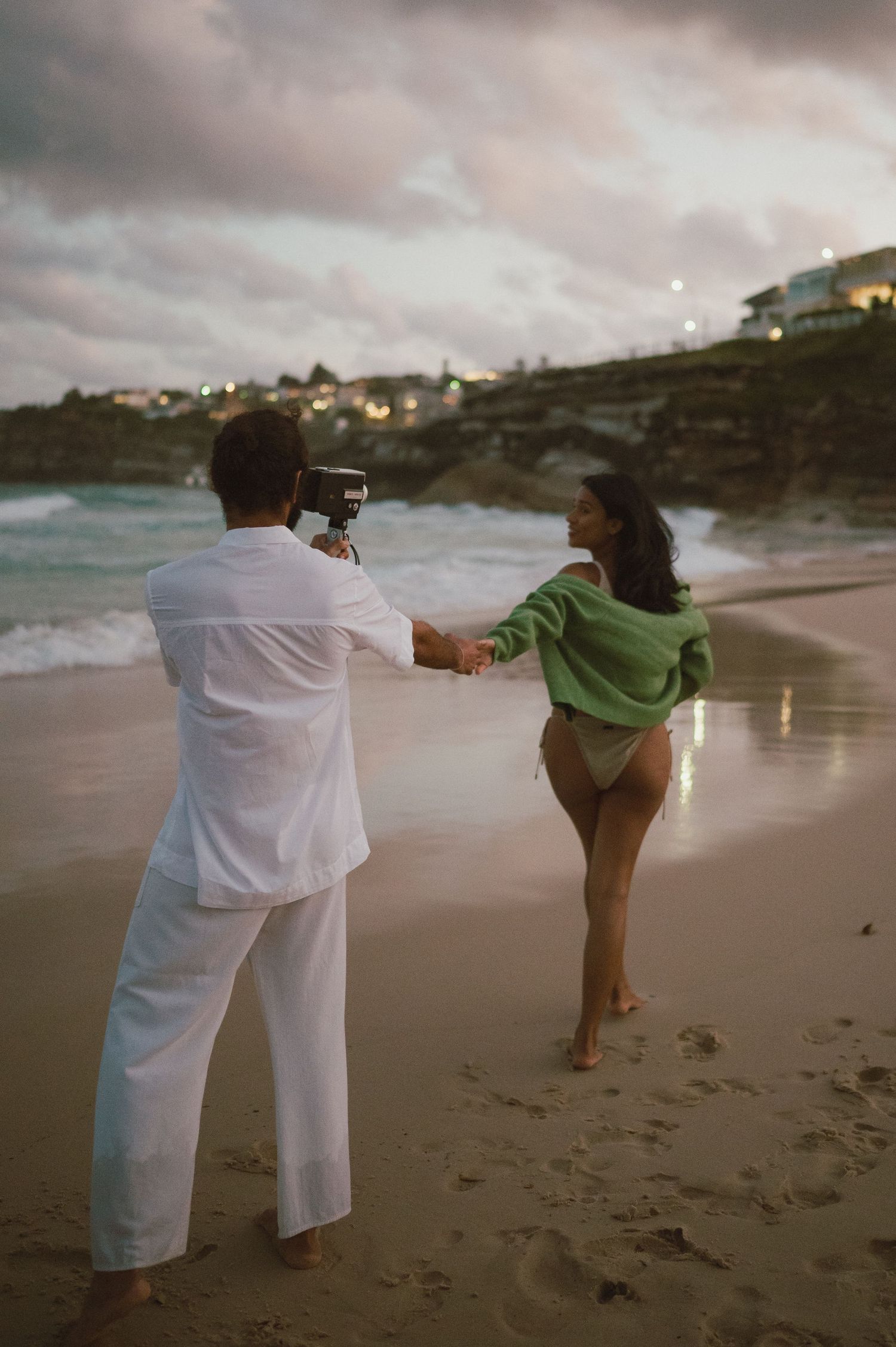 Two people on a beach at dusk, one taking photos while the other poses.