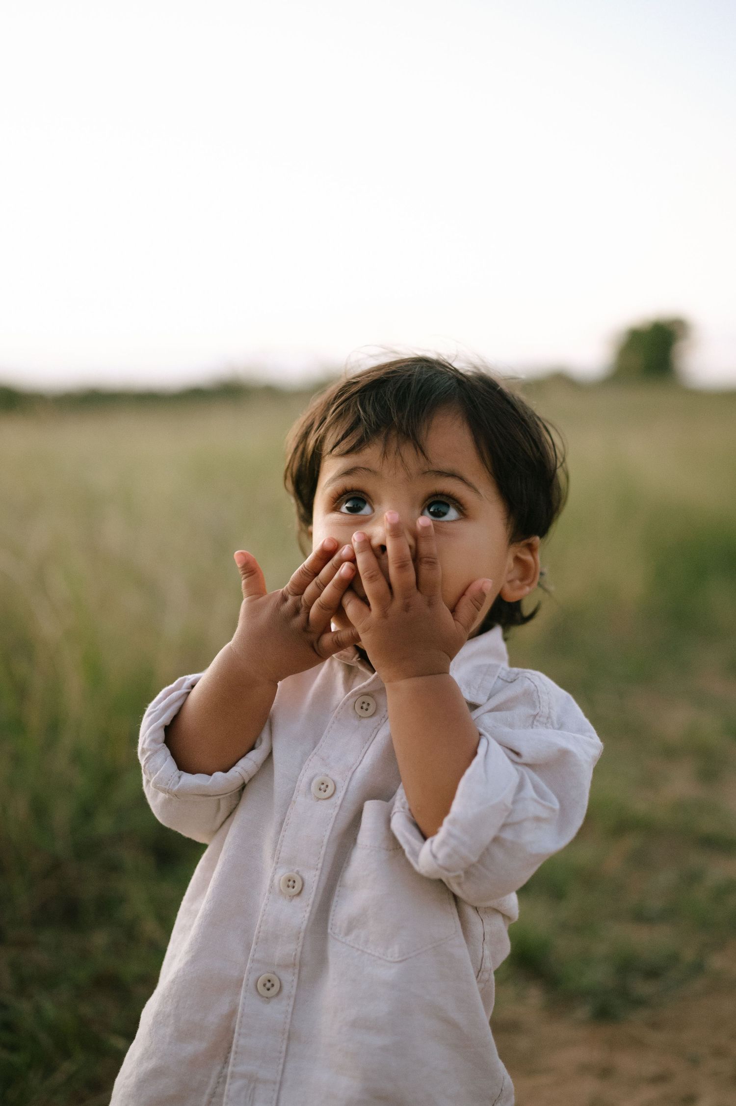 A toddler in a white shirt makes playful expressions while sitting in a grassy field.