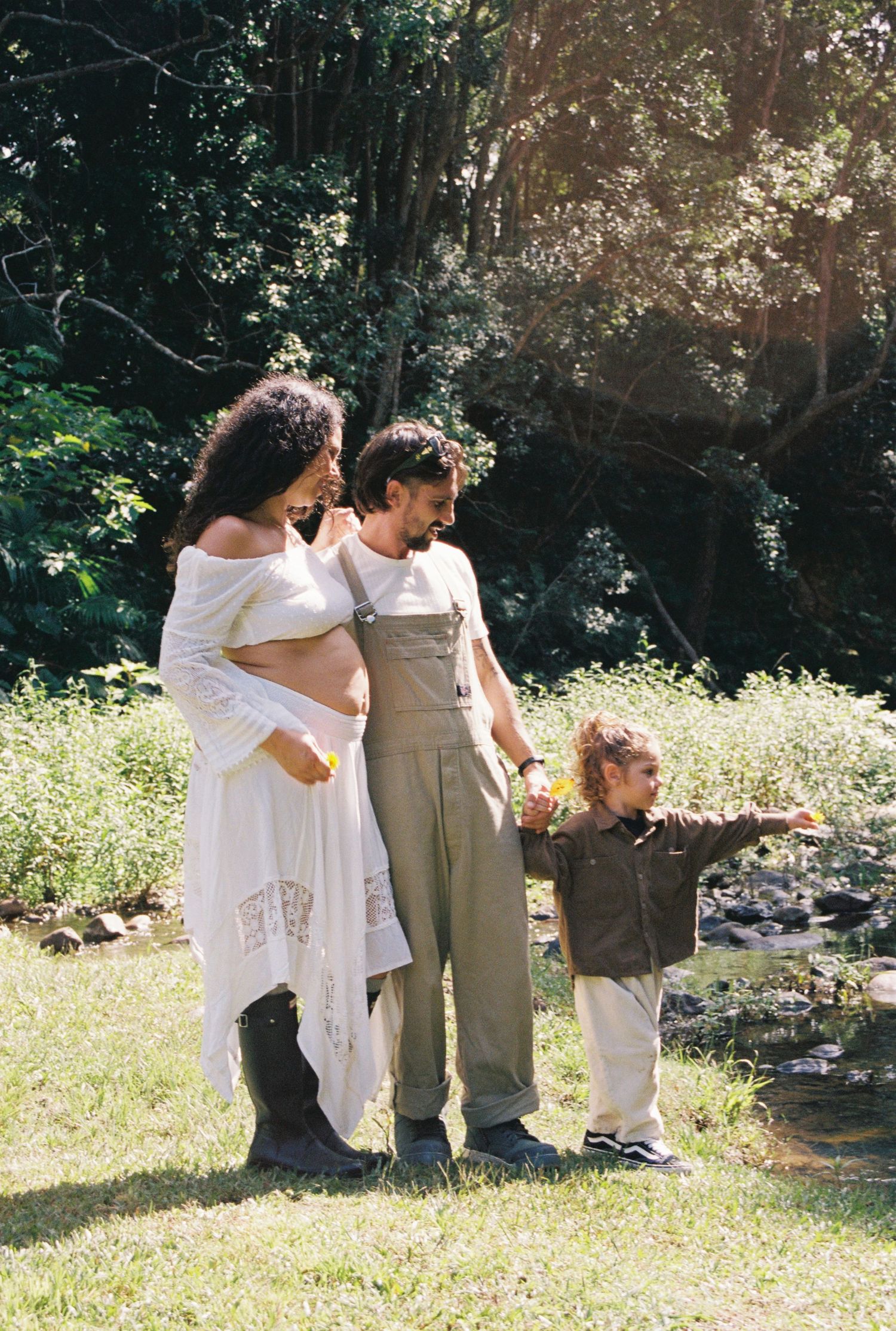 Pregnant woman in white dress with man in overalls and toddler pointing outdoors in lush green park setting.