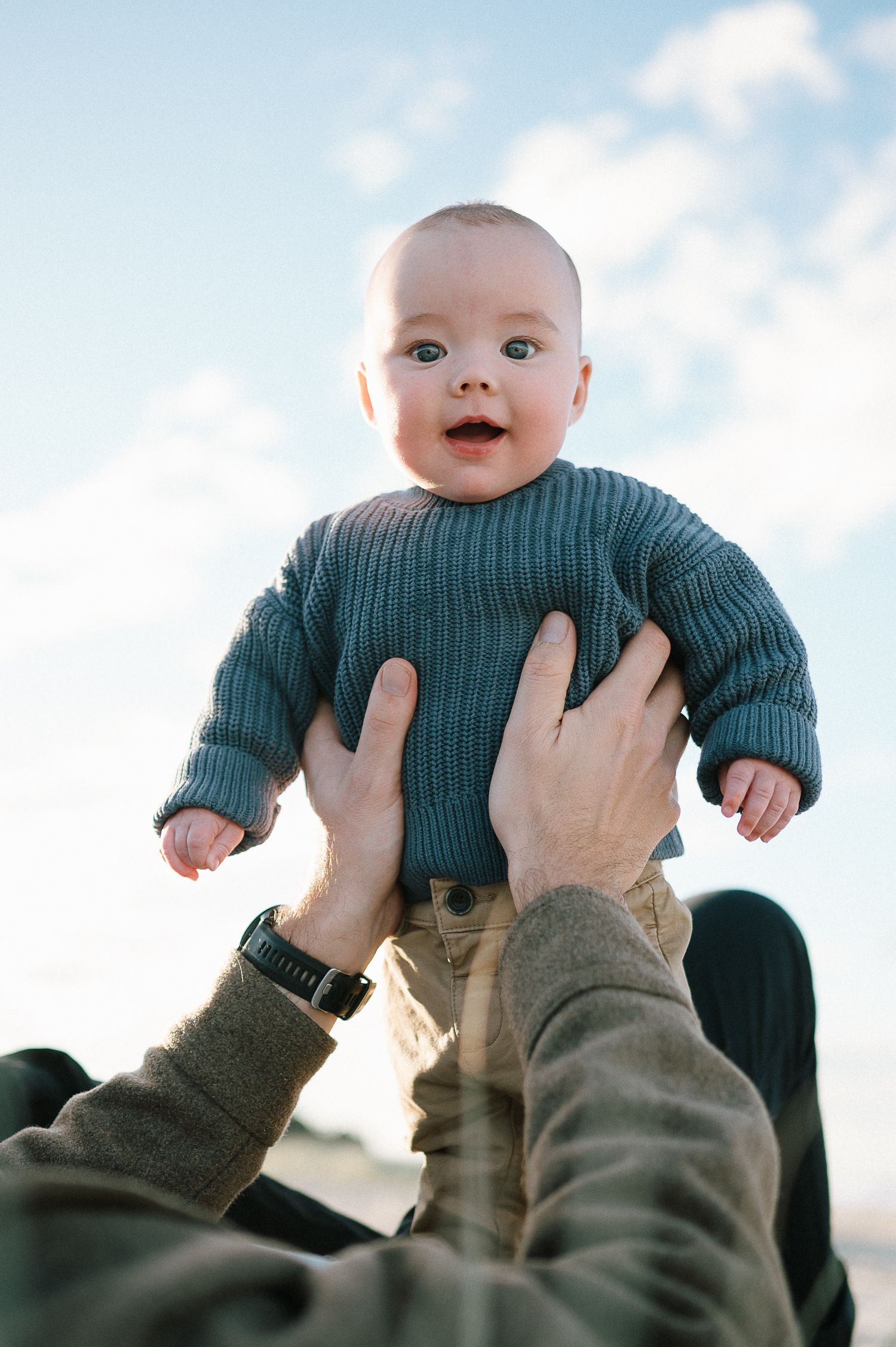 Baby in teal sweater being lifted up against blue sky background.