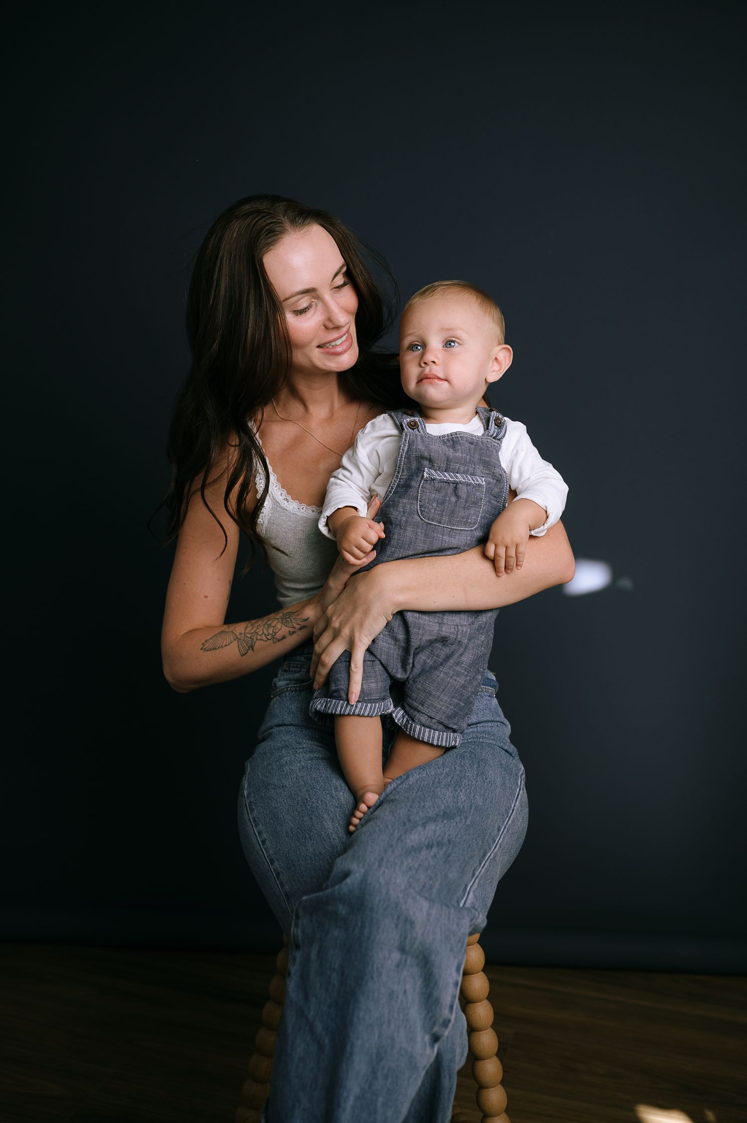 A smiling mother with dark hair holds her baby in denim overalls against a dark studio background.