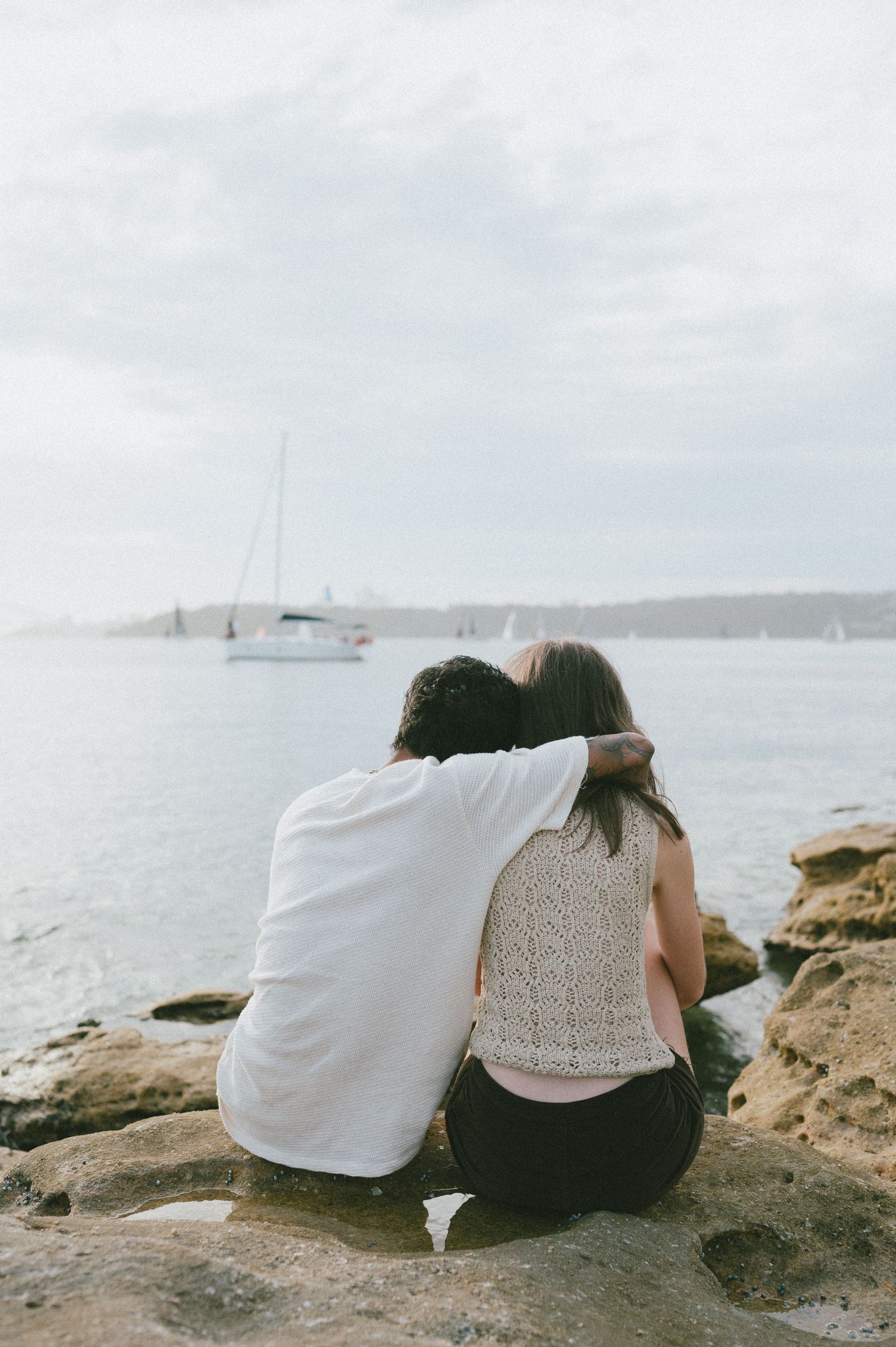 A couple sits close together on coastal rocks overlooking a harbor view.