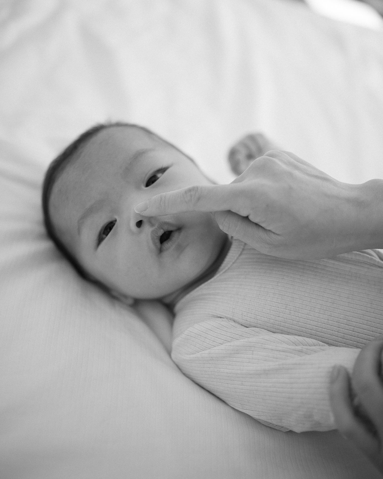 Black and white photo of a newborn baby lying on white sheets while an adult gently touches its nose.