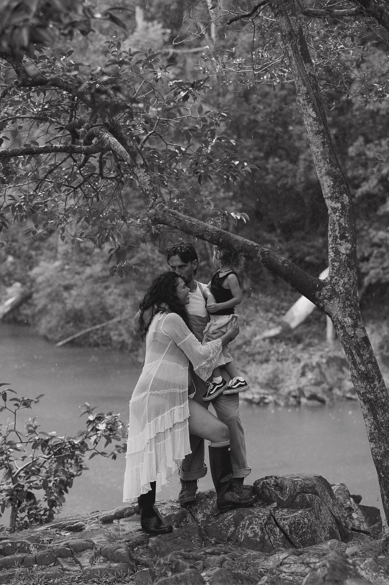 Black and white photo of a couple sharing a romantic moment with a child by a river surrounded by trees.