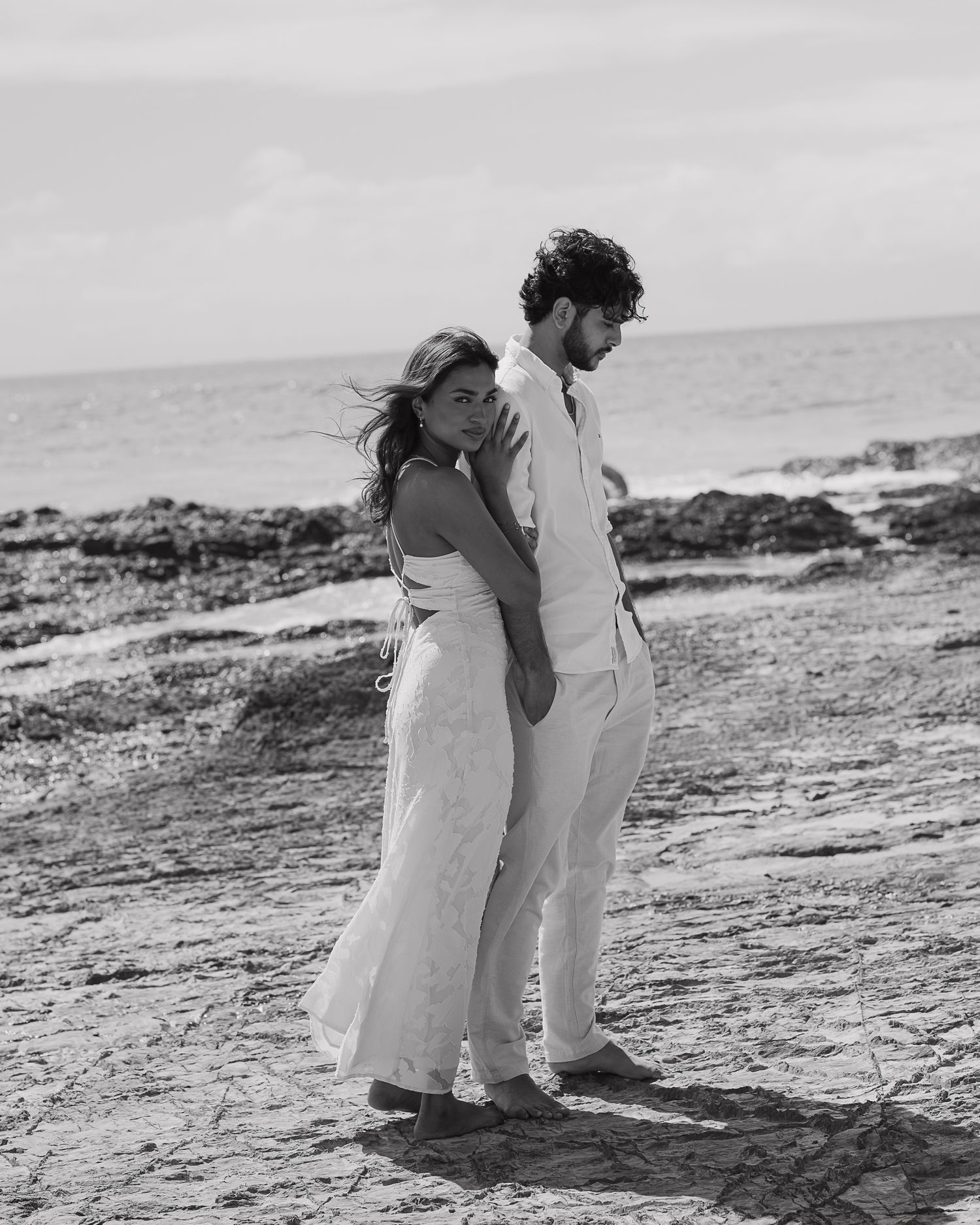 A couple embraces on a rocky beach shoreline in black and white.