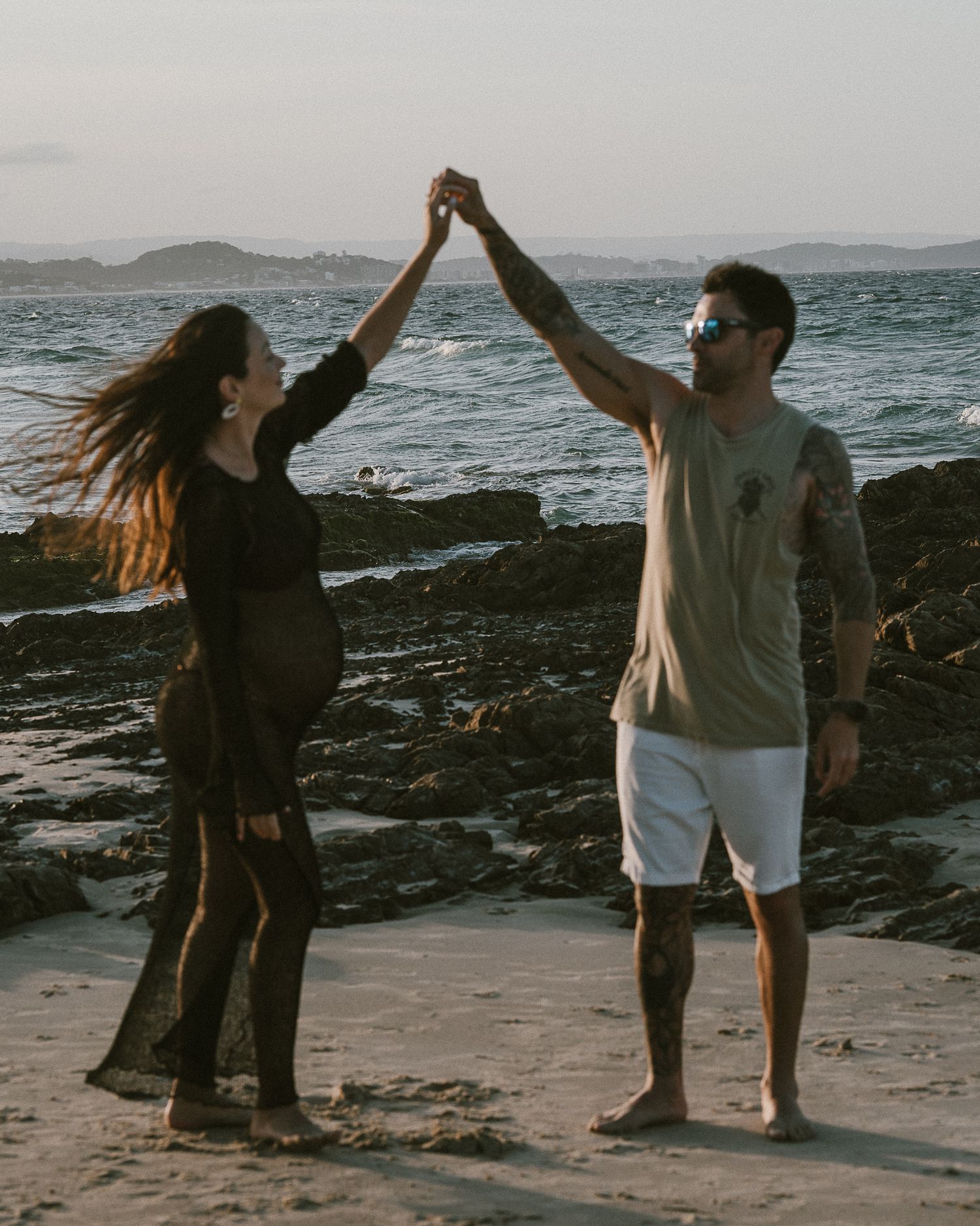 Couple dancing on a rocky beach at sunset, holding hands raised, woman in black dress, man in white shorts.