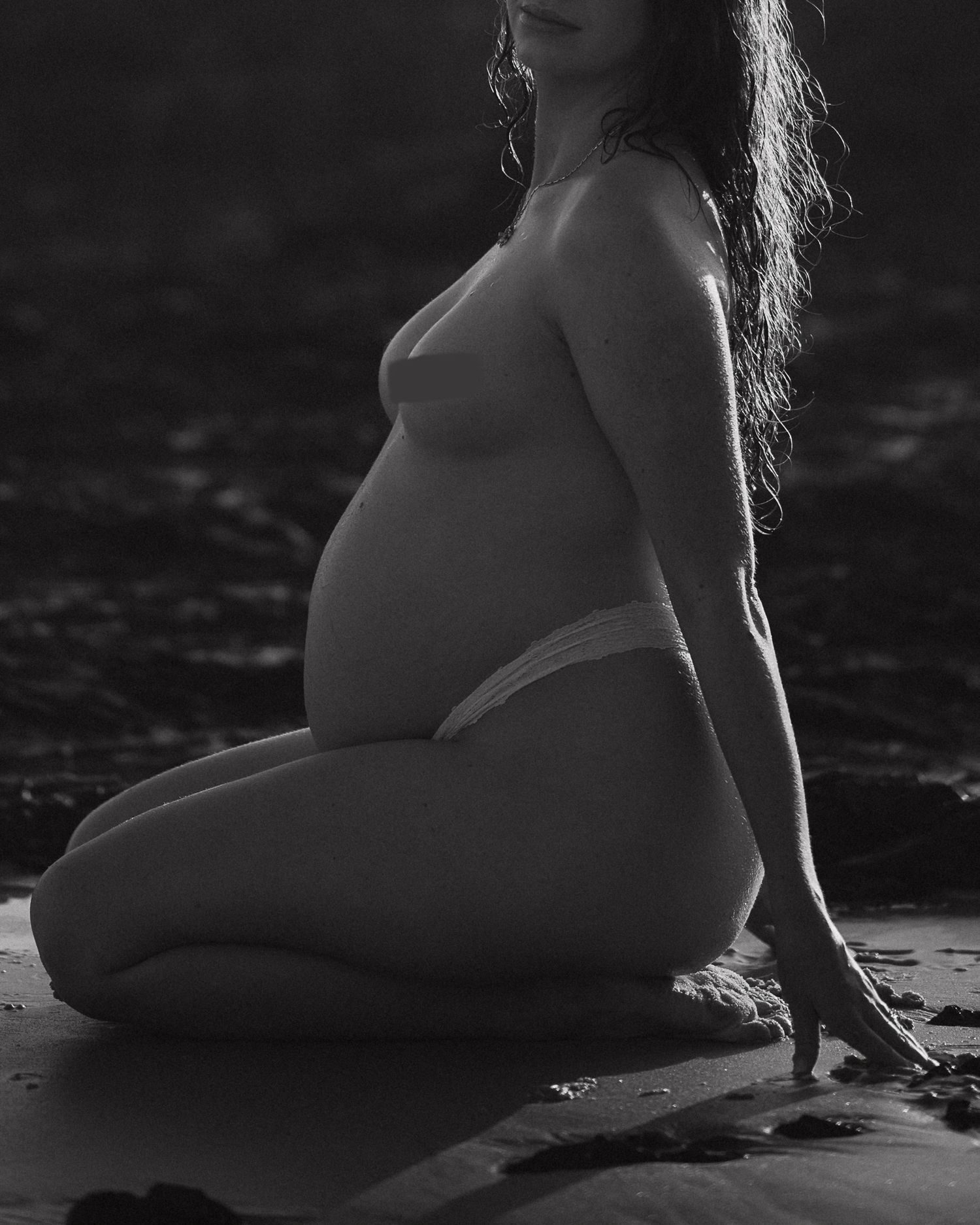 Dramatic black and white maternity photo showing pregnant figure reclining on beach.
