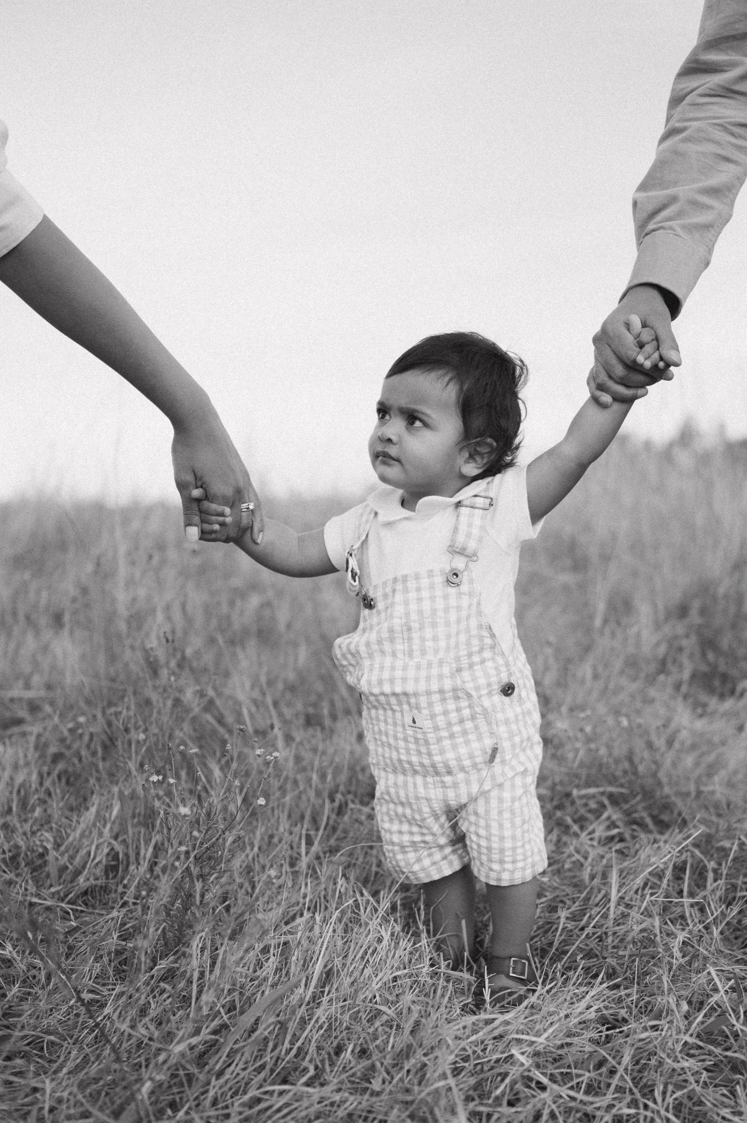 Black and white photo of toddler in checkered overalls holding hands with adults in field.
