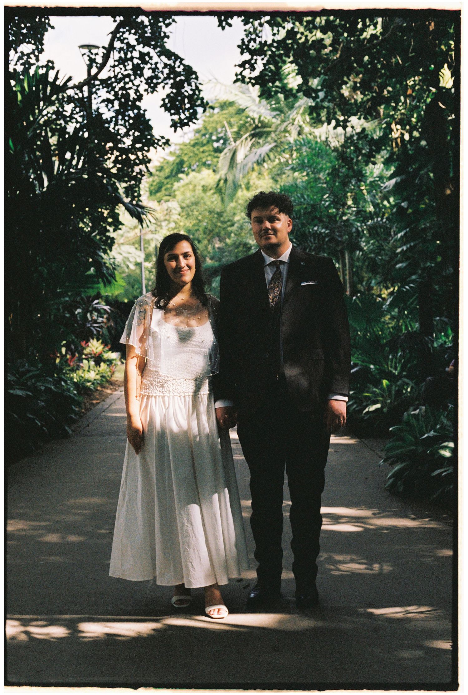 Couple in wedding attire walking along a lush garden pathway, bride in white dress, groom in dark suit.
