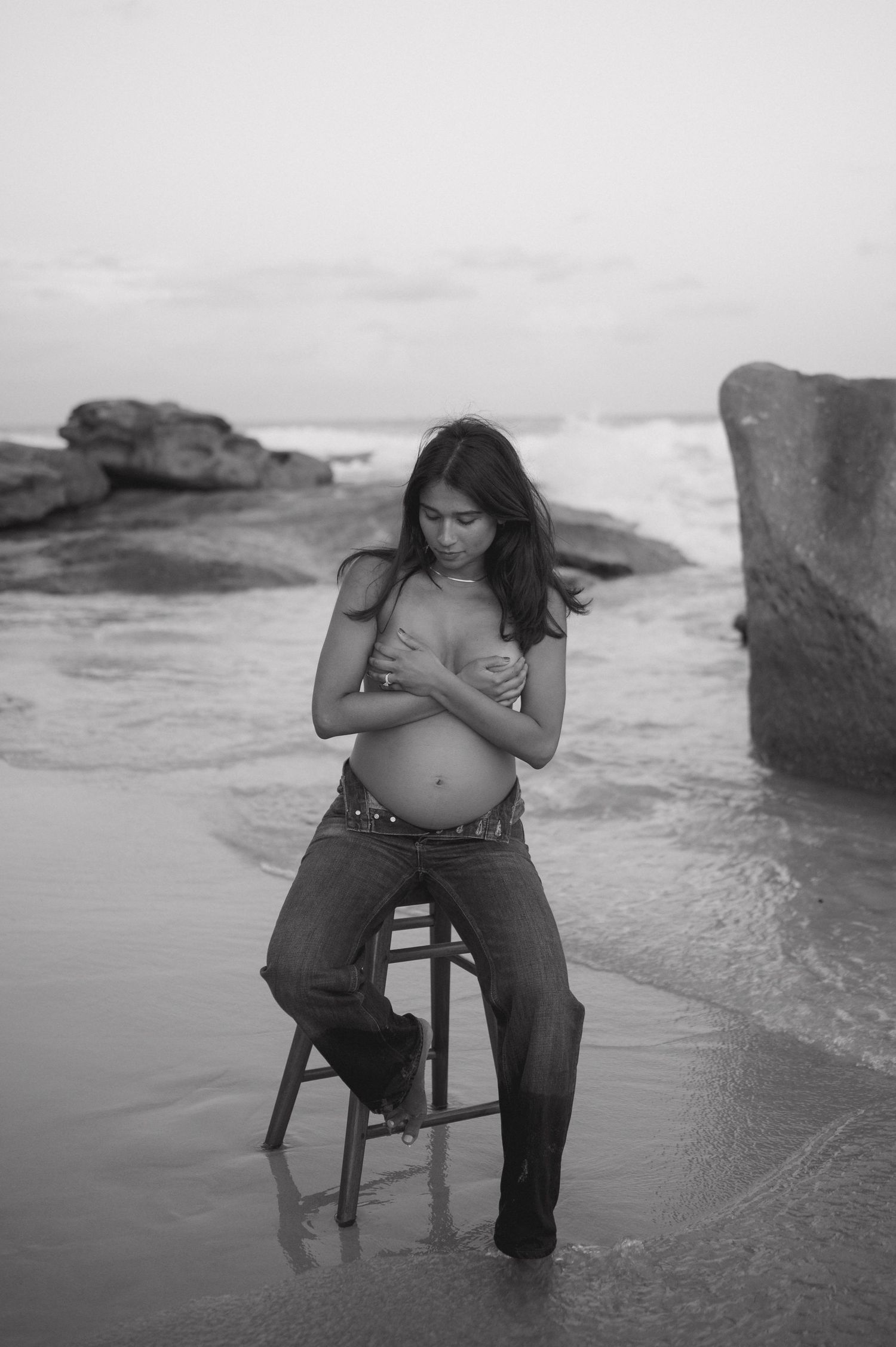Black and white beach maternity session with wooden chair in shallow surf.