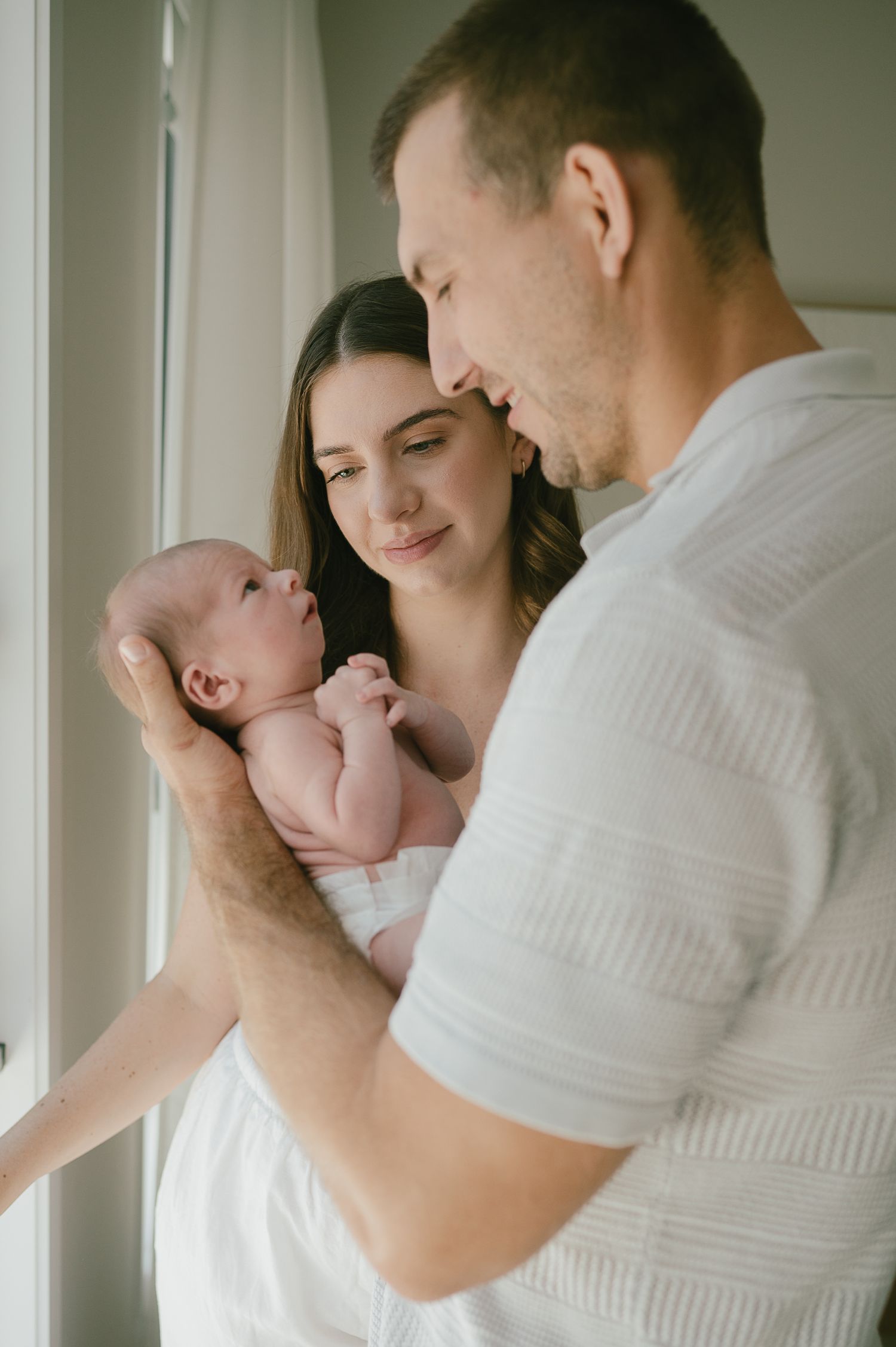 A couple tenderly gazes at their newborn baby near a bright window in a soft, intimate family portrait.