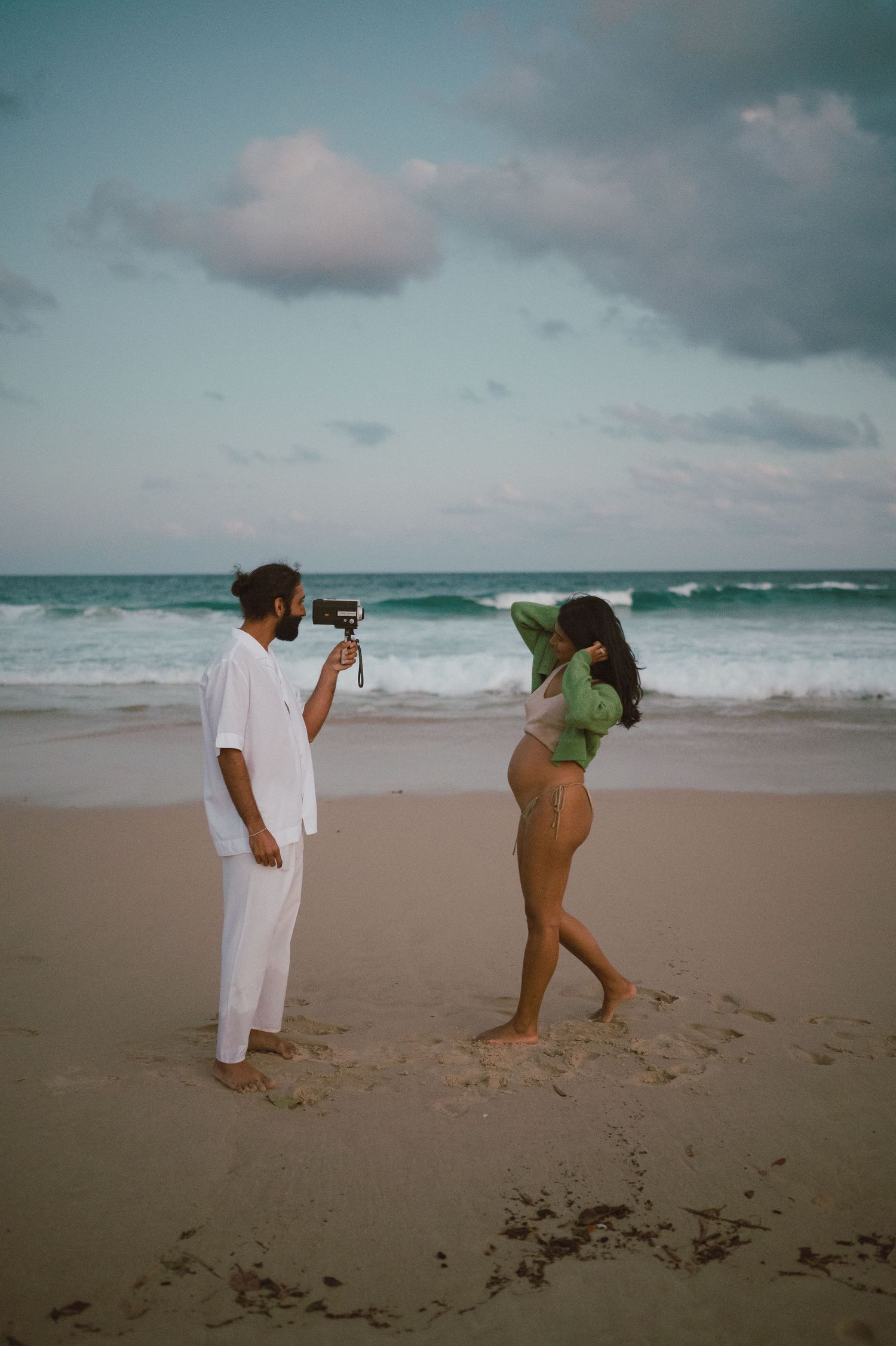 Couple playfully interacting on a sandy beach with crashing waves and cloudy skies in the background.