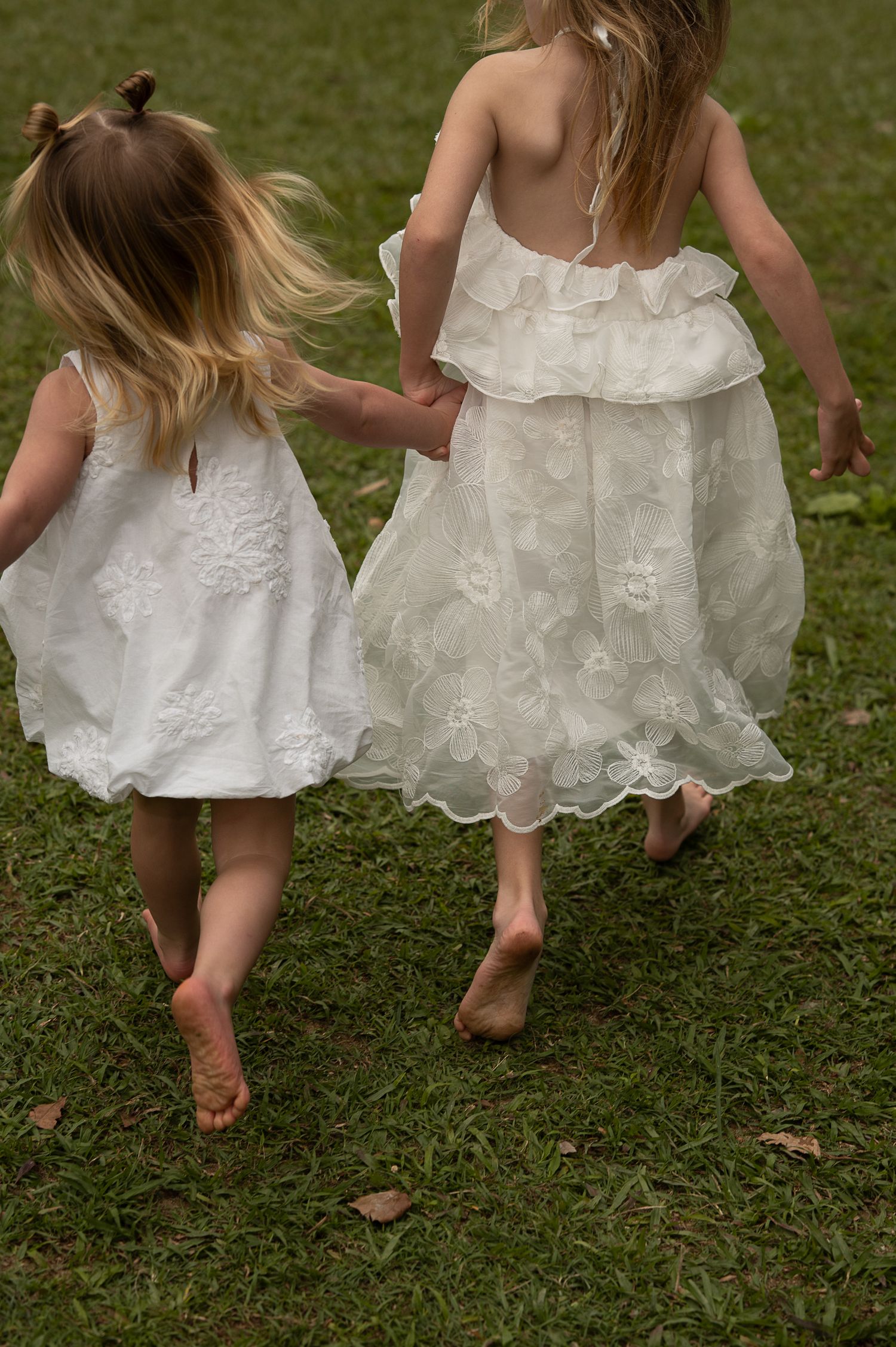 Two young girls in white lace dresses hold hands while running barefoot on green grass.