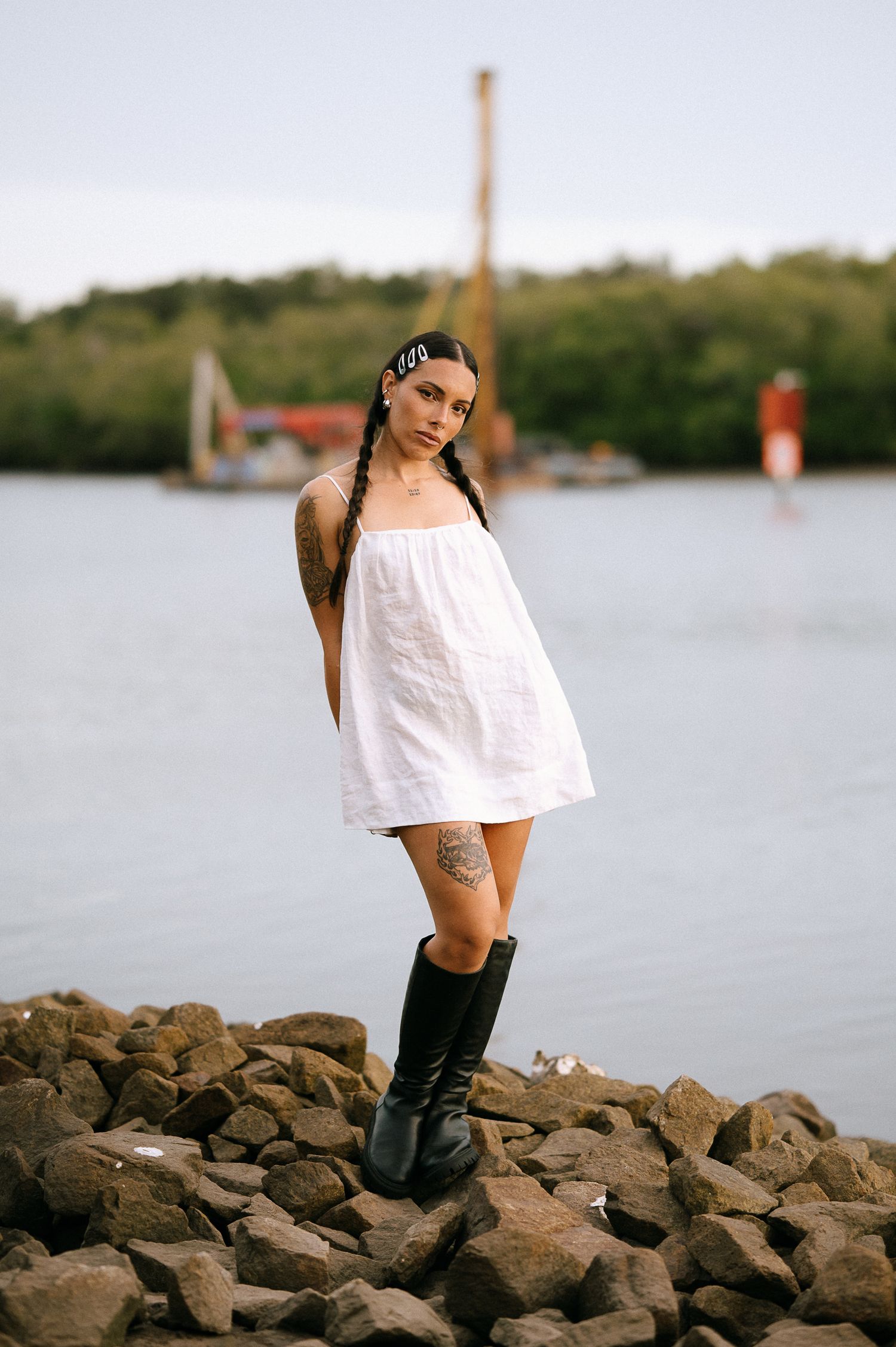 Figure in white dress and boots standing on rocky shoreline with lighthouse in background.